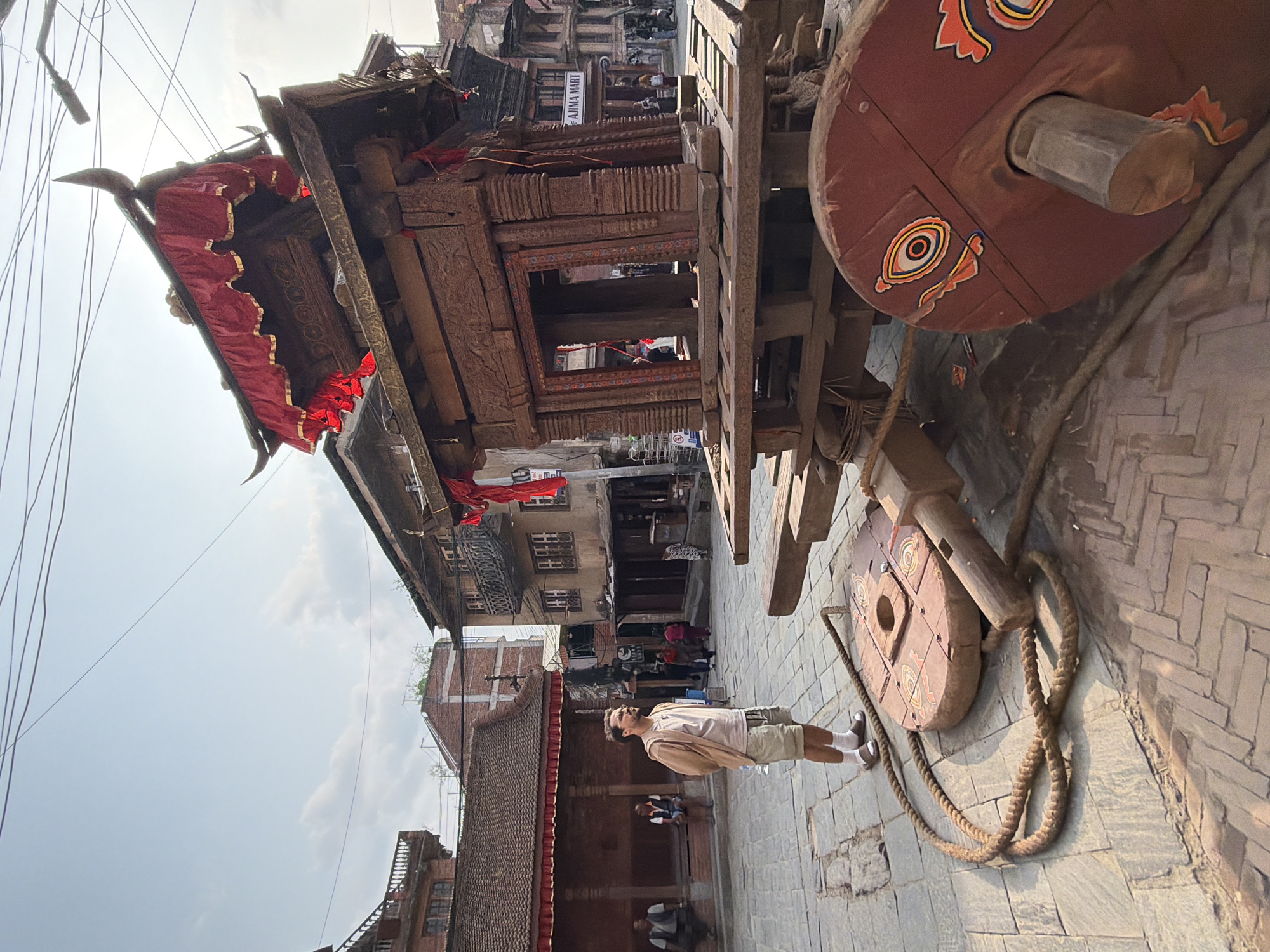 The image captures a scene in a historic town, featuring a person observing a large, ornate wooden structure, likely a ceremonial chariot or rath. The perspective is slightly upward, with a bright, partly cloudy sky visible at the top.
In the foreground, a man stands on a paved street, looking up and towards the large wooden structure. He has a beard and is dressed in a light-colored sleeveless shirt layered over a light brown long-sleeved shirt, light-colored shorts, white socks, and dark shoes.
To the right of the man, the prominent wooden structure dominates a significant part of the frame. It is intricately carved with traditional designs, showcasing rich dark wood. Red fabric banners with gold trim are draped from its upper sections. The structure is supported by massive wooden wheels; one in the foreground is painted red with distinctive circular, eye-like motifs in yellow, black, and white. Thick, coiled ropes lie on the stone and brick paved ground next to the chariot's wheels.
The background reveals traditional multi-story buildings with brick walls and characteristic wooden window frames and balconies, indicative of classic Newari architecture. A sign on one of these buildings displays the text "AJIMA MART." The street is paved with a mix of stone slabs and brick patterns. The bright sunlight suggests it is daytime, possibly late morning or early afternoon. The scene conveys a sense of cultural immersion and admiration for historical artifacts and architecture in a vibrant, old-world setting.