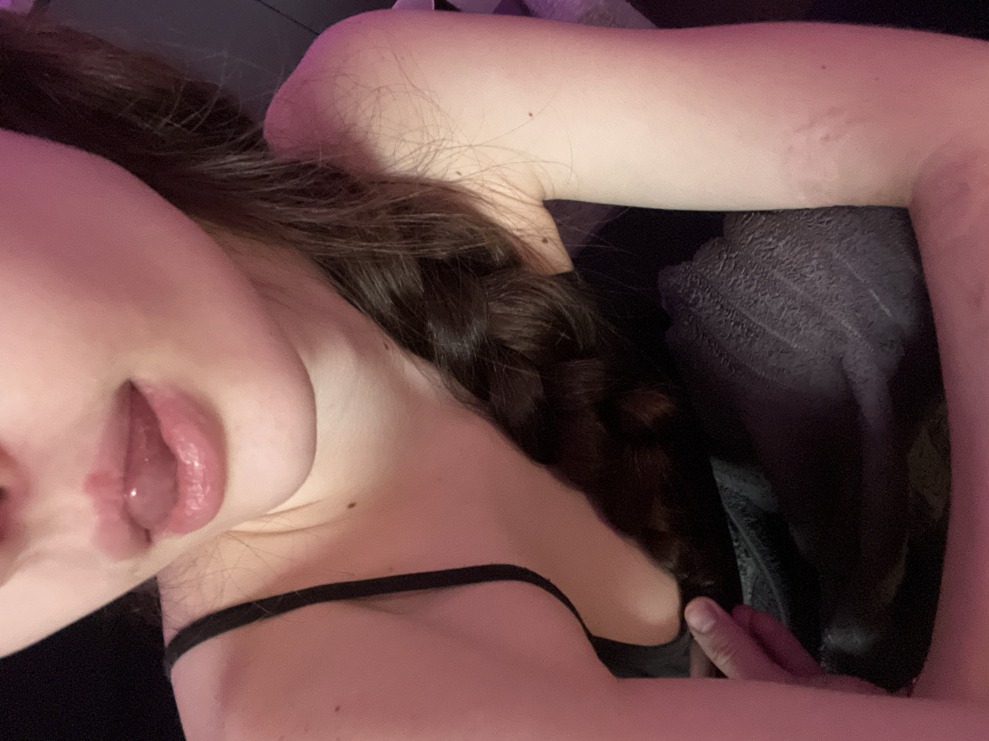 A close-up, angled shot depicts a person from the chest up. The individual has fair skin, dark braided hair, and is wearing a black top with thin straps. Their lips are parted, revealing a glimpse of their tongue. The person's right arm is raised, partially obscuring the face. The background is dark and blurry, showing textured fabric. The lighting is dim and pink-toned. Location: Unknown, Russia.