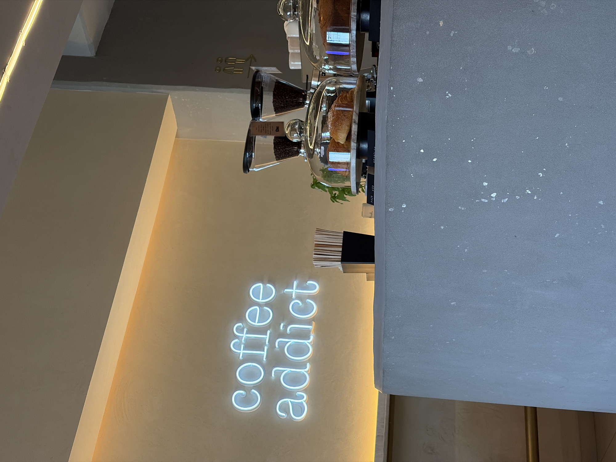 The image captures the interior of a modern coffee shop or cafe, featuring a minimalist design with a neutral color palette. The scene is dominated by a display counter and a warmly lit wall.
On the left, a light beige-toned wall is illuminated by a radiant light blue neon sign that spells out "coffee addict" in a casual script. A thin strip of warm white lighting runs along the top edge of the wall, enhancing the ambiance. Further up and to the left, architectural details of the ceiling and an upper dark grey beam are visible, along with what appears to be metal wall fixtures or sconces.
To the right, a sturdy counter with a textured, matte grey surface, possibly concrete or a similar composite material flecked with lighter specks, runs across the foreground. On the counter, several items are arranged: two clear glass cloches covering freshly baked golden-brown croissants, and two conical glass dispensers filled with dark coffee beans, each with small labels. A small green plant sits near the base of one of the pastry cloches, adding a touch of freshness. A black container holding white straws or stirrers is also present on the counter. The overall setting suggests a contemporary and inviting space.