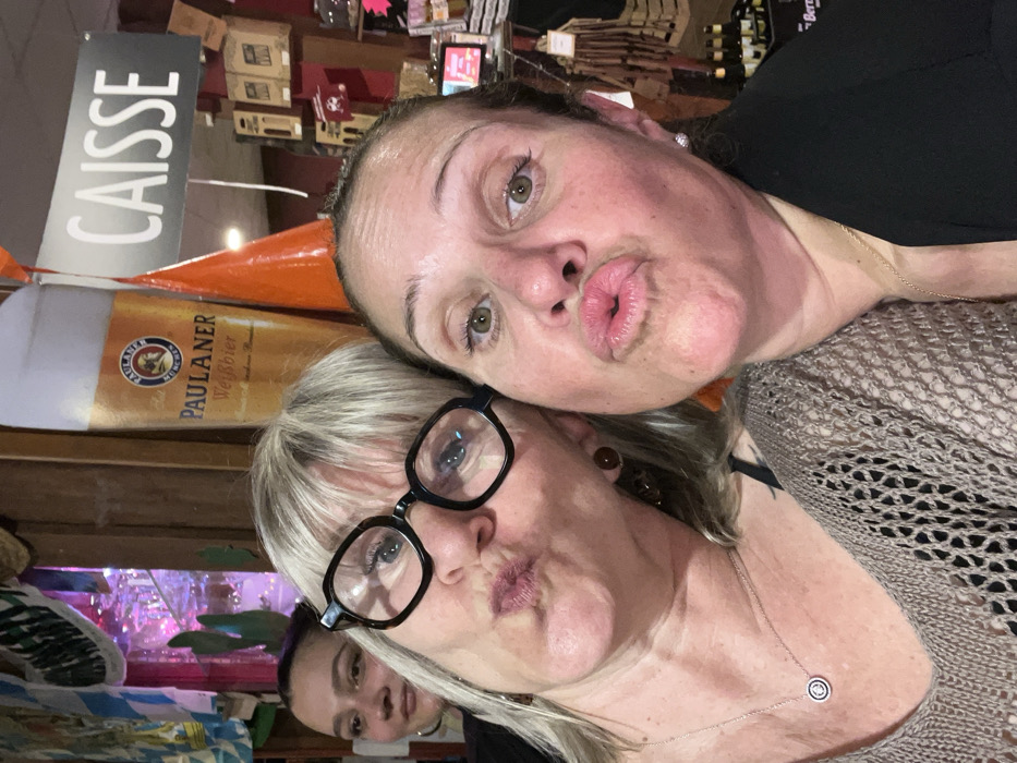 A close-up photograph captures three women in an indoor setting, identified as a retail establishment or bar in L'Étrat, France. Two women occupy the foreground, positioning themselves for the camera with puckered lips. The woman on the left has blonde hair, wears black-framed glasses, brown circular earrings, and a silver necklace with a circular pendant, dressed in a beige open-knit top. The woman on the right has darker hair, visible silver stud earrings, and wears a black top or blazer. A third woman with dark hair is partially visible in the left background, smiling subtly. The background features a large cardboard cutout of a Paulaner Weißbier glass. Above it hangs a gray sign with "CAISSE" (French for "cash register") in white capital letters, attached to an orange conical object. Shelving filled with various products, including bottles, is visible in the background, alongside decorative elements such as blue and white patterned fabric and green cactus-shaped items. The overall lighting is warm, with some purple illumination present in the far left background.