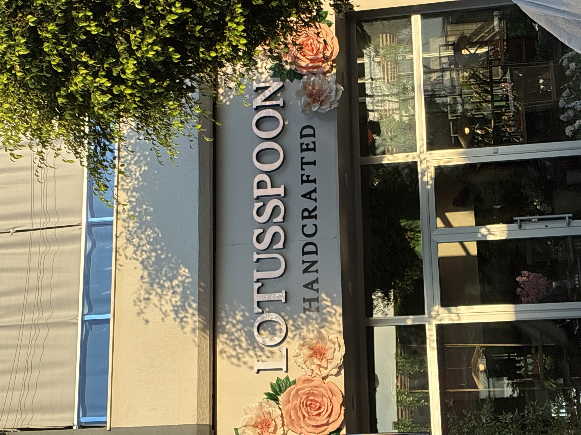 The image shows the storefront of "LOTUSSPOON HANDCRAFTED" in Alanya, Turkey. The facade is a light beige color, with the store name displayed in large, metallic letters. Decorative fabric flowers in shades of pink and peach are affixed to the wall around the signage. Below the signage are large glass windows and doors, revealing the interior of the shop which appears to be a boutique or gift shop, with displays of various items including what seem to be bottles, vases, and decorative objects. A large green tree with vibrant leaves partially obscures the right side of the building and signage, casting dappled shadows across the facade. The time of day appears to be late afternoon or early evening, as indicated by the warm lighting and the long shadows. The sky above is a muted grey, partially covered by a grey awning.