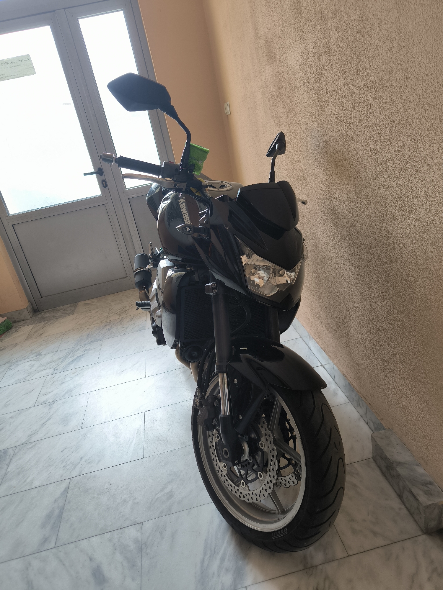 A black Kawasaki motorcycle is parked indoors against a peach-colored wall. The floor is tiled with large, light-colored marble tiles. A white door with glass panels is visible in the background. The specific location within Sofia, Bulgaria cannot be determined from the image alone.