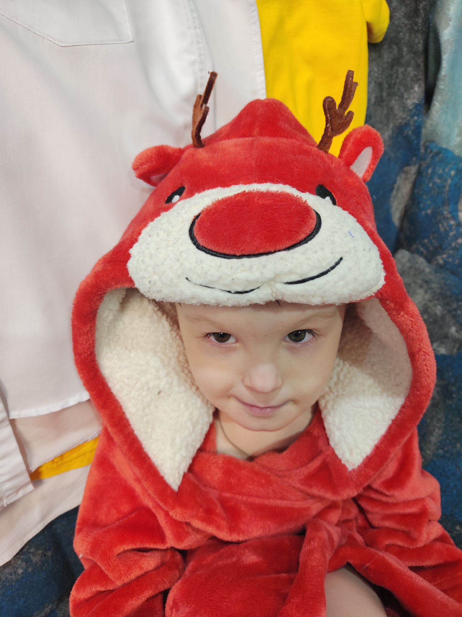 A young child is wearing a red reindeer hooded blanket. The child has light skin, light brown hair, and brown eyes. The hood has a large, round, red nose, white fur trim, and small brown antlers. The child is looking at the camera with a slight smile. Behind the child, there is a white shirt, a yellow shirt, and a blue and gray blanket. The location is Chaikovsky, Russia.
