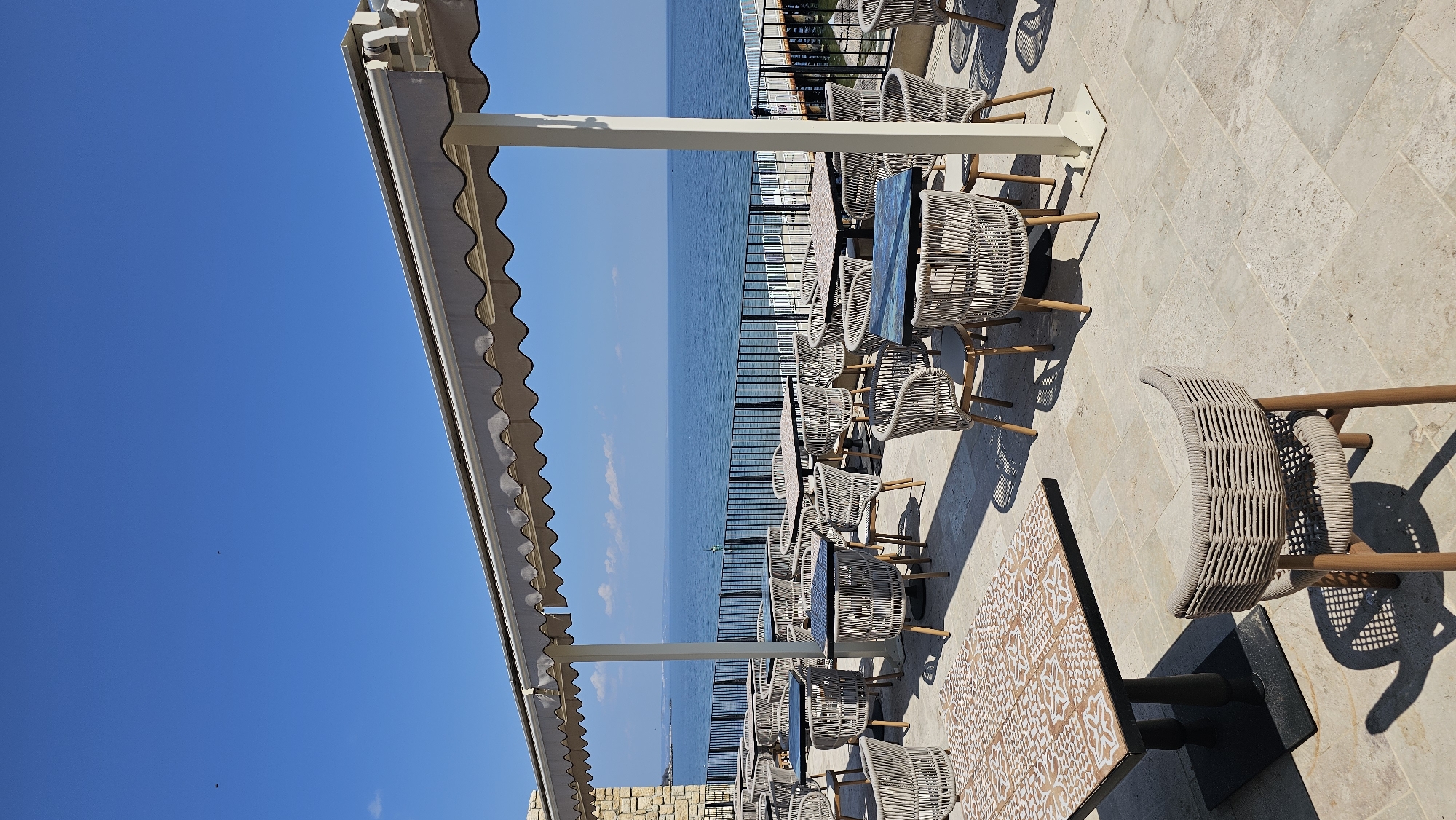 The image depicts an outdoor dining area likely in Varna, Bulgaria, given the view of the Black Sea. The scene is set on a stone-paved patio with several tables and chairs. The chairs are made of a light-colored woven material with wooden legs. Tables have a variety of surfaces, including plain and patterned designs. A light-colored awning provides shade for the dining area, supported by white posts. In the background, a black metal railing separates the patio from the sea. The sea is visible in the distance, with a clear blue sky above. The image appears to be taken during the day, with strong sunlight casting shadows from the furniture. There are no people visible in the scene.
