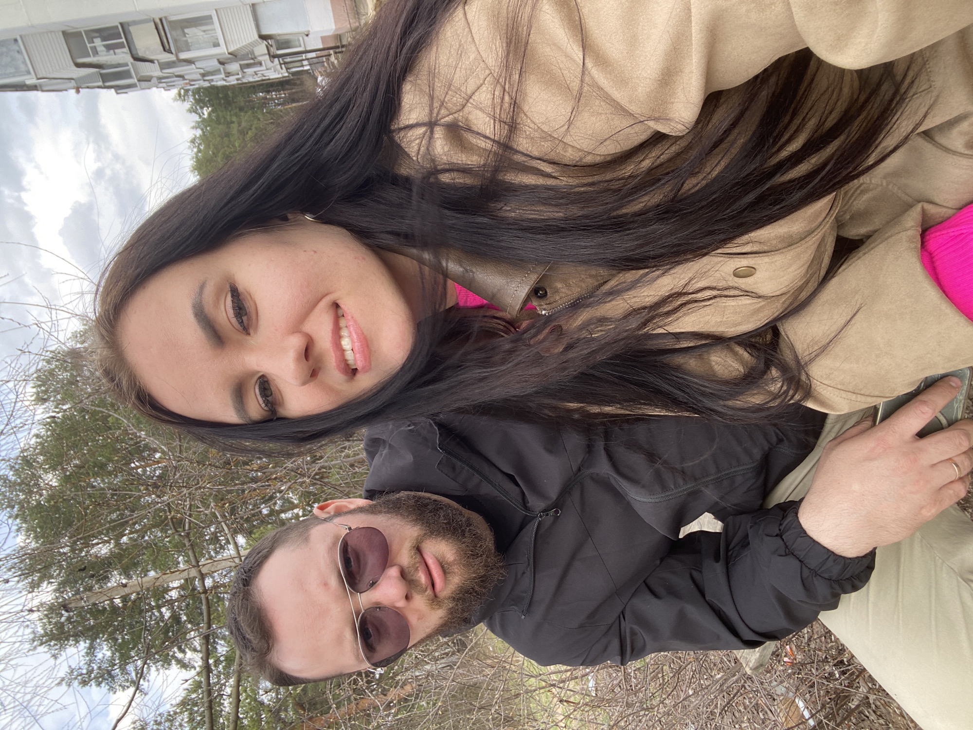 This image captures a selfie of two individuals, a woman and a man, taken outdoors in Chaikovsky, Russia. The perspective is from slightly above and angled, with both subjects looking towards the camera.
The woman is positioned on the right side of the frame, smiling broadly with her teeth visible. She has long, dark brown hair flowing over her shoulders and chest. She is wearing a light tan or beige jacket over a bright fuchsia pink top. Her eyes are dark, and she has visible eyeliner and mascara, with a gold hoop earring visible on her right ear.
On the left side of the frame is the man, looking directly at the camera with a neutral, relaxed expression. He has a full beard and mustache and is wearing sunglasses with reddish-brown lenses. He is dressed in a black zip-up jacket and light tan or khaki pants. His right hand, adorned with a gold band on the ring finger, is visible near his lap, holding a small, dark object that appears to be a phone or similar device.
The background features bare branches of trees or shrubs in the immediate foreground, suggesting late winter or early spring. Behind them, a large, multi-story building with numerous windows, likely an apartment complex, is visible under a cloudy sky. Patches of green trees can be seen further in the distance. The overall weather appears cool and overcast, indicating daytime. The individuals are positioned close together, suggesting a friendly or intimate relationship. No visible text is present in the image.