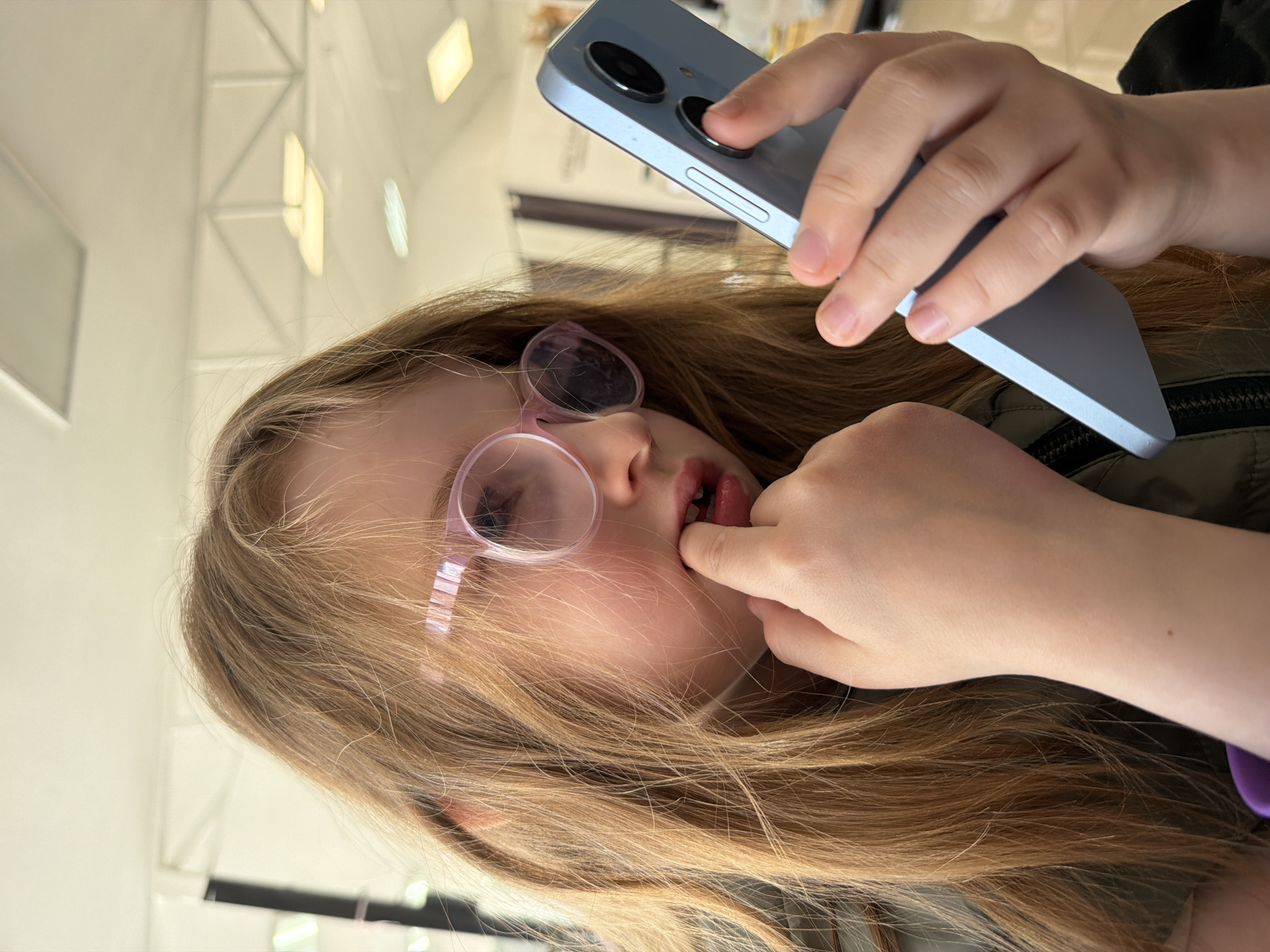 The image features a young girl, likely a child or pre-teen, with long, light brown hair, wearing round, light pink-rimmed glasses and a dark green zipped jacket. She is positioned with her head tilted, and her left hand is raised, with a finger touching her lips or teeth, indicating concentration or a habitual gesture. In her right hand, she holds a light blue smartphone, its back visible with two distinct circular camera lenses and a side button. Her gaze appears to be directed towards the phone screen, which is not visible.
The setting is indoors, characterized by a bright, light-colored ceiling with several rectangular artificial lights, some of which are illuminated. Structural elements, possibly a ceiling truss or architectural beams, are also visible in the background, suggesting a large public space. The artificial lighting dominates the scene, making the time of day undeterminable. Given the location context of Chaikovsky, Russia, this could be a shopping center, public building, or transport hub. There is no visible text in the image. The girl's expression suggests focus or contemplation while interacting with her phone. The overall image is rotated 90 degrees counter-clockwise.