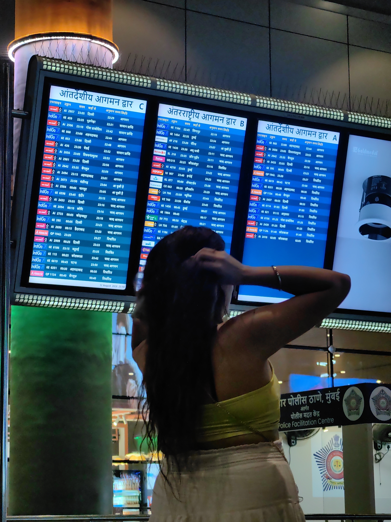 The image shows a digital flight information display board at an airport. The board displays arrivals and departures, with flight numbers, destinations, and times. The text on the board is in Hindi and English. In the foreground, a woman is seen from behind, with her hands in her hair. She is wearing a yellow top and a light-colored skirt. The background shows airport infrastructure, including lights and signs. A prominent sign in the background reads "Police Facilitation Centre" in English and "पुलिस मदत केंद्र" in Hindi, indicating that the location is in Mumbai, India. The date and time displayed on the flight board is "12 August 2023 23:10".