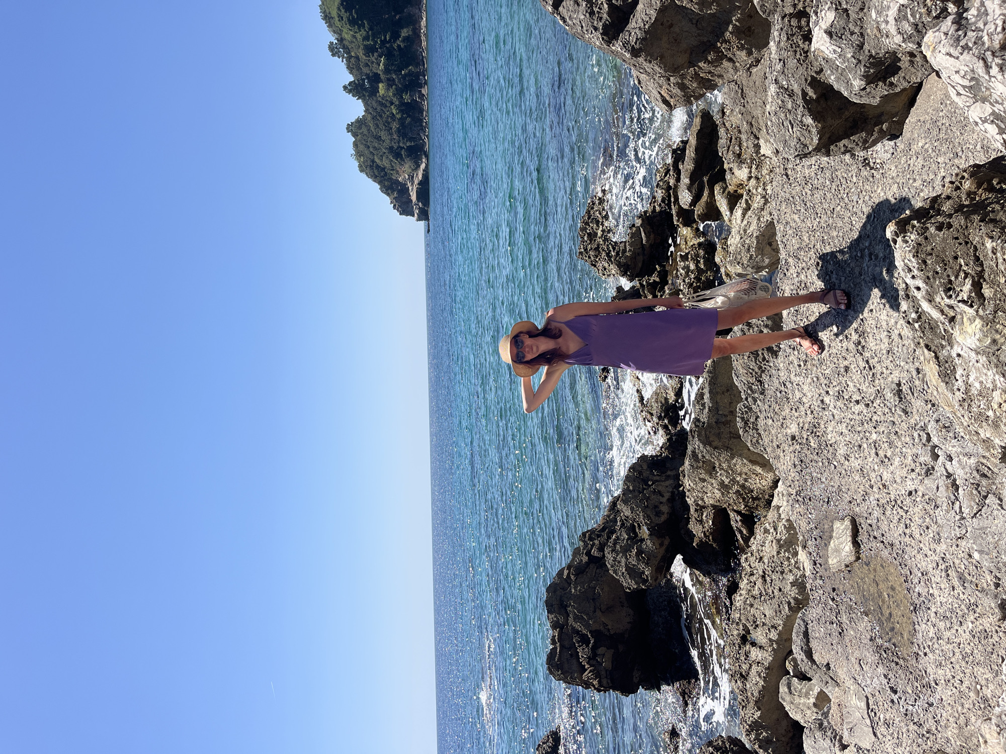 A woman wearing a purple dress and a straw hat stands on a rocky outcrop next to the sea. The sea is blue and stretches to the horizon. A headland covered in trees is visible in the distance. The sky is clear and blue. The location is possibly near Budva, Montenegro, given the coastal landscape and architectural style.