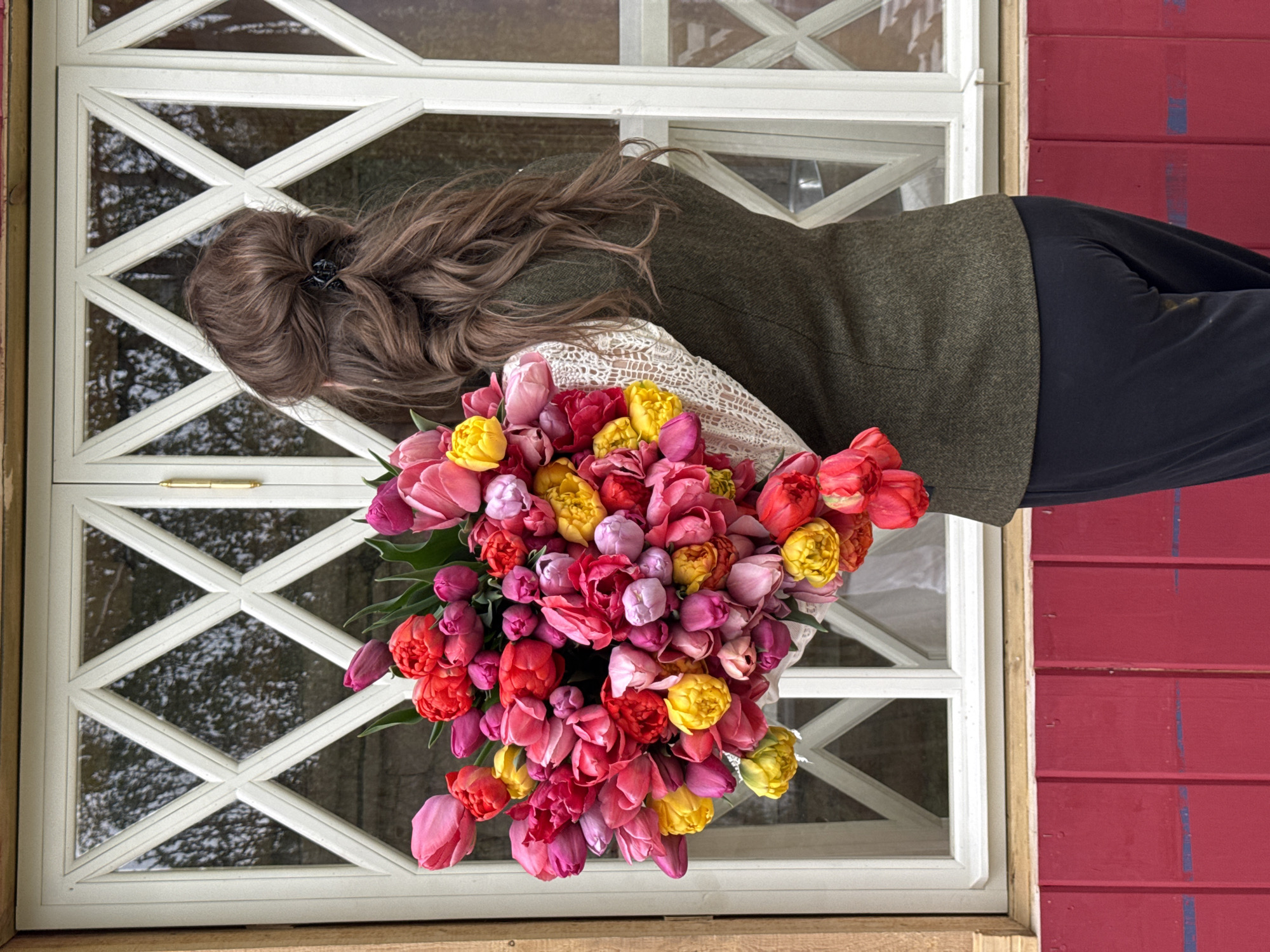 A woman with long brown hair, partly held up by a clip, stands in front of a window with a white frame and a patterned glass. She wears a green jacket over a white lace top and black trousers. She holds a large bouquet of colorful tulips, primarily in shades of red, pink, and yellow. The setting appears to be outdoors, with a red wooden wall adjacent to the window. The photograph was taken in Sapronovo, Russia.
