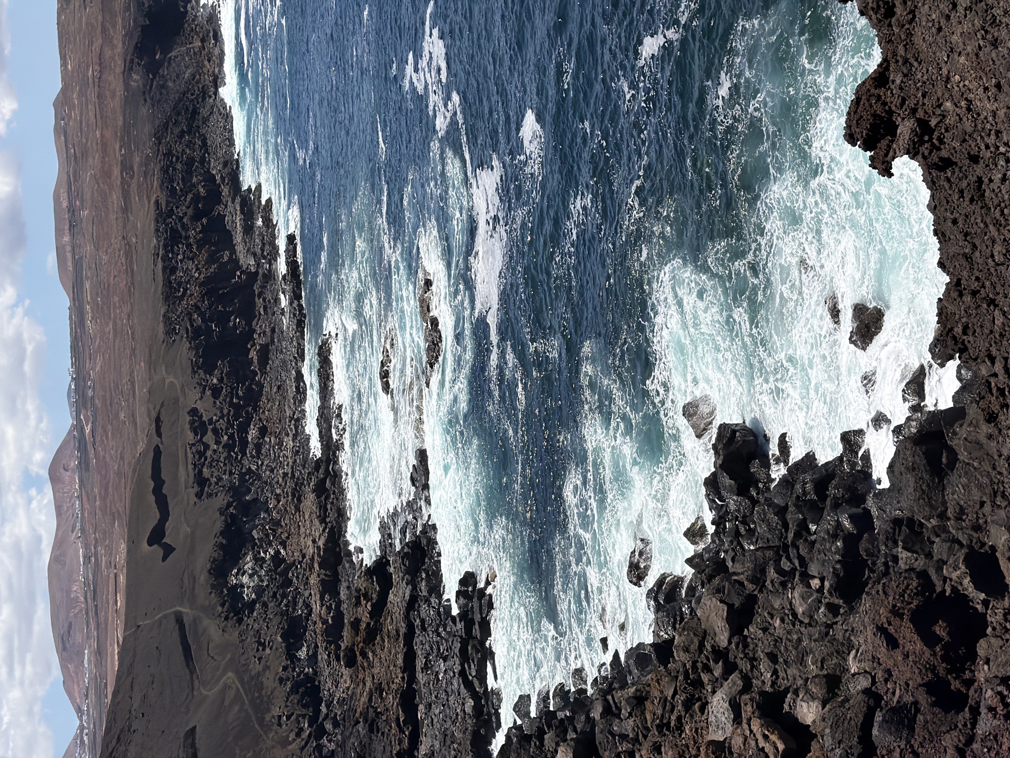 The image shows a coastline of volcanic rock in Tinajo, Spain, with crashing waves. The dark, jagged rocks form a stark contrast against the turquoise and white foam of the Atlantic Ocean. In the background, a brown, rocky landscape extends towards the horizon under a blue sky.