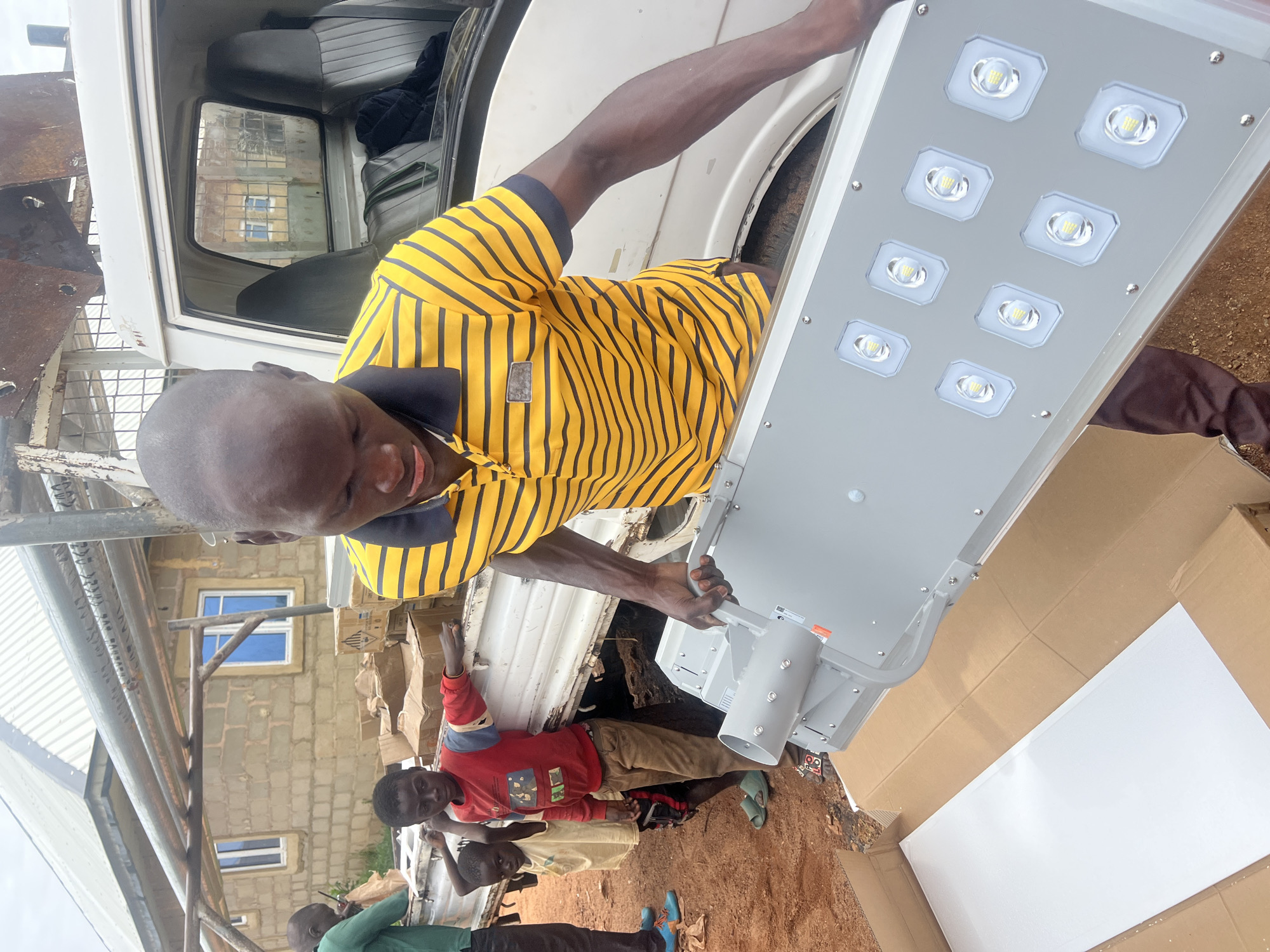 The image depicts an outdoor scene with a man in a yellow and black striped shirt holding a solar-powered street lamp. He is standing next to a white truck that has its bed loaded with cardboard boxes. Two young boys are also present, standing near the truck bed. One boy is wearing a red shirt and the other a yellow shirt. In the background, a building constructed with concrete blocks and a metal roof is visible. The ground appears to be dirt. The overall setting suggests a delivery or installation of solar lighting equipment in a rural or semi-urban area, possibly in Dong, Nigeria, given the context. The man is in the process of handling the lamp, which has multiple LED lights.