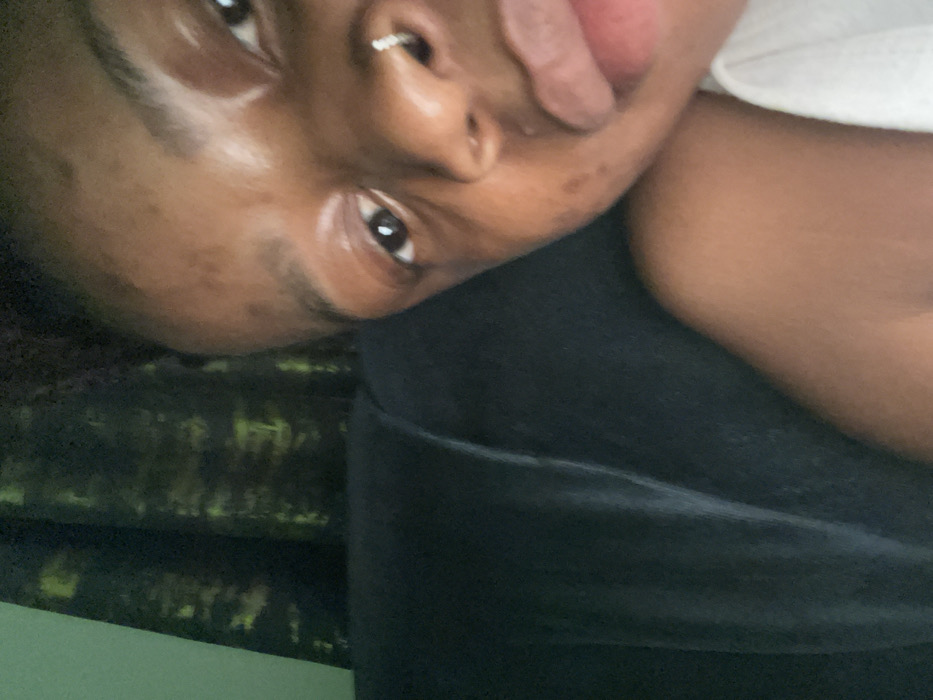 A person with smooth, brown skin and a nose piercing leans close to the camera. Their lips are slightly parted. One eye is visible and appears dark. The person is wearing black clothing and resting against a couch with a patterned, green and gold upholstery. A hint of a white shirt is seen near their neck.