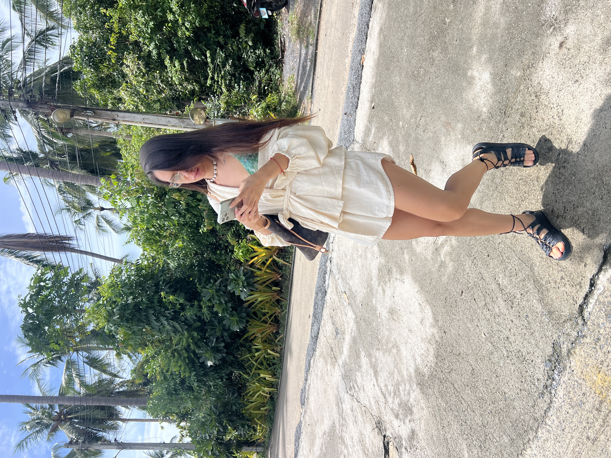 A woman with long dark hair and glasses is standing on a paved road, looking at her phone. She is wearing a white top with a tie closure at the waist, a green bikini top underneath, white shorts, and black lace-up sandals. She has a dark handbag slung over her shoulder. The background consists of lush green trees, palm trees, and a bright blue sky with scattered clouds. There are power lines visible above the trees. To the right, a motorcycle is parked on the side of the road. The scene appears to be in a tropical or sub-tropical environment.