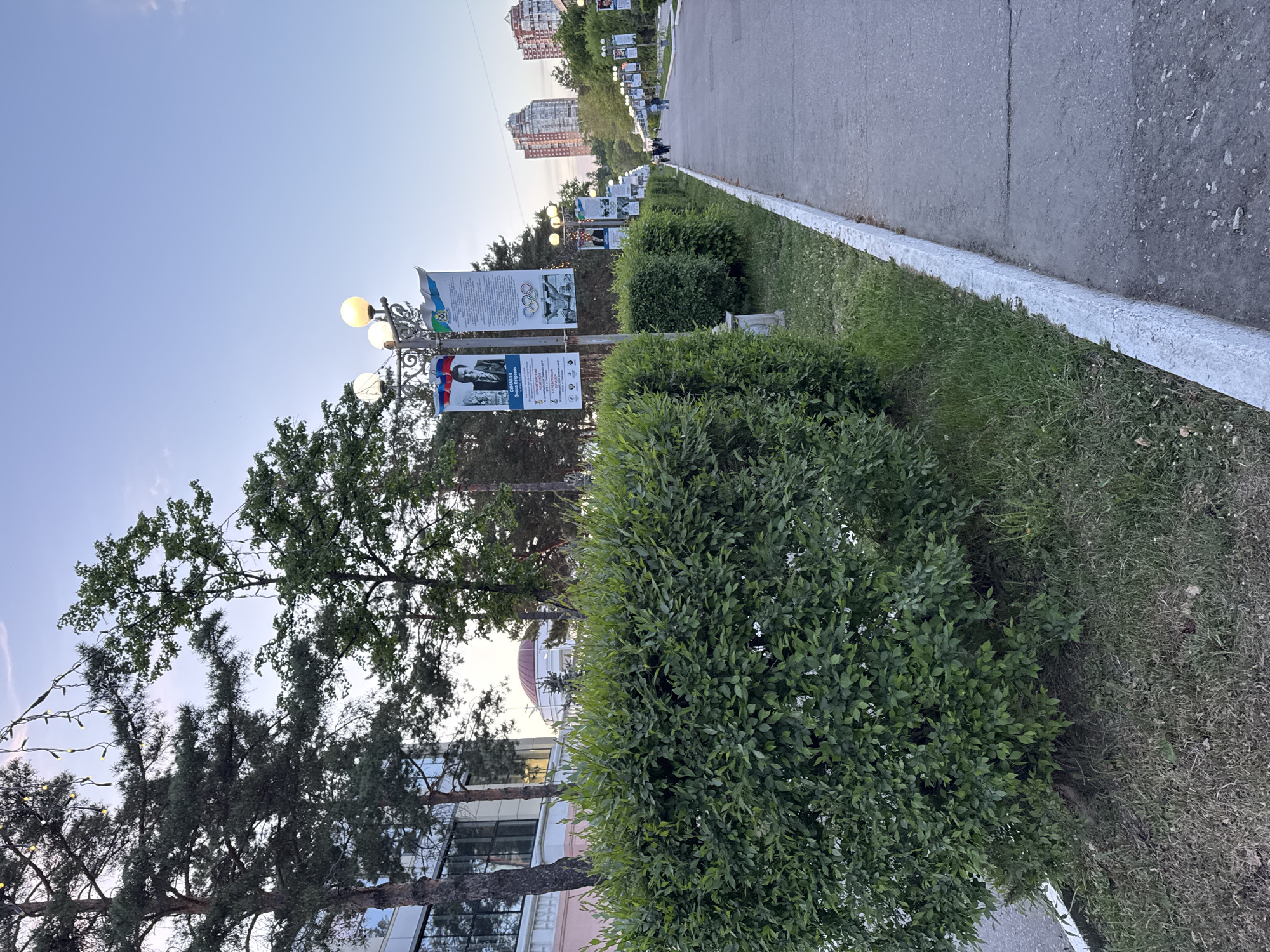 The image depicts a sidewalk in the city of Khabarovsk, Russia. Along the sidewalk, there are bushes, trees, and lampposts. The lampposts have historical information banners attached to them. In the background, there are apartment buildings and a few people walking. The sky is pale blue. The sidewalk is made of asphalt and has a white curb.