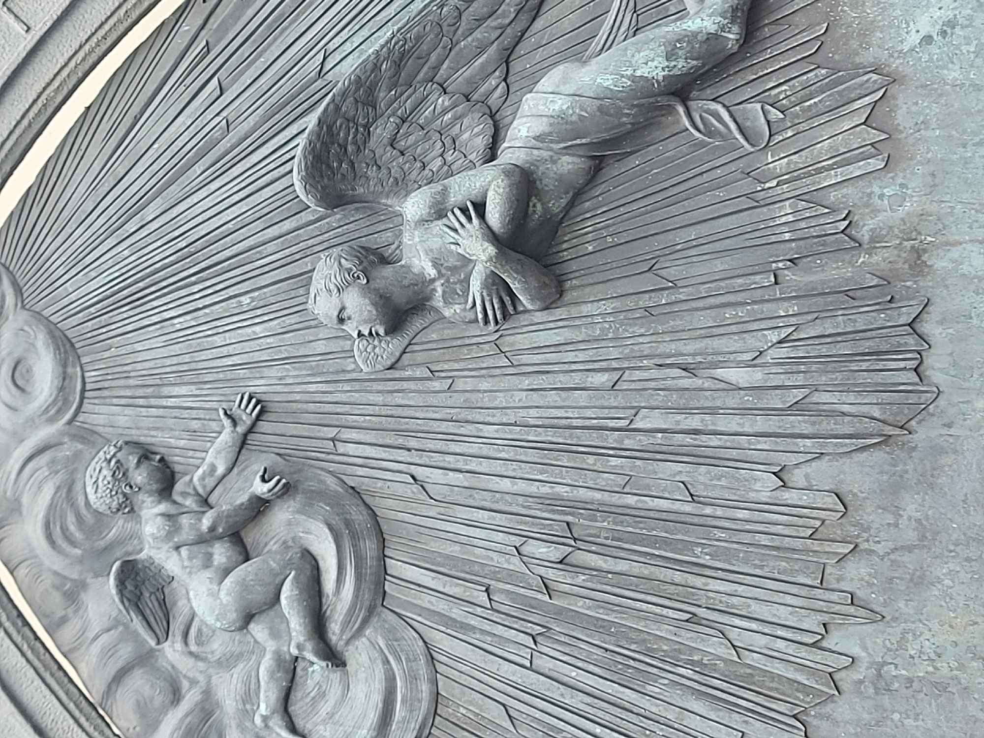 The image shows a bronze or similar metallic relief sculpture depicting two angels. One angel, positioned in the upper right, is depicted in a recumbent pose, with wings extended and hands clasped. The second angel is smaller and located on the lower left, emerging from a cloud formation with arms raised. The background consists of radial lines that radiate outwards from the cloud towards the center, creating a sense of divine light or energy. The metallic surface has a weathered or oxidized appearance. The location is St. Petersburg, Russia.