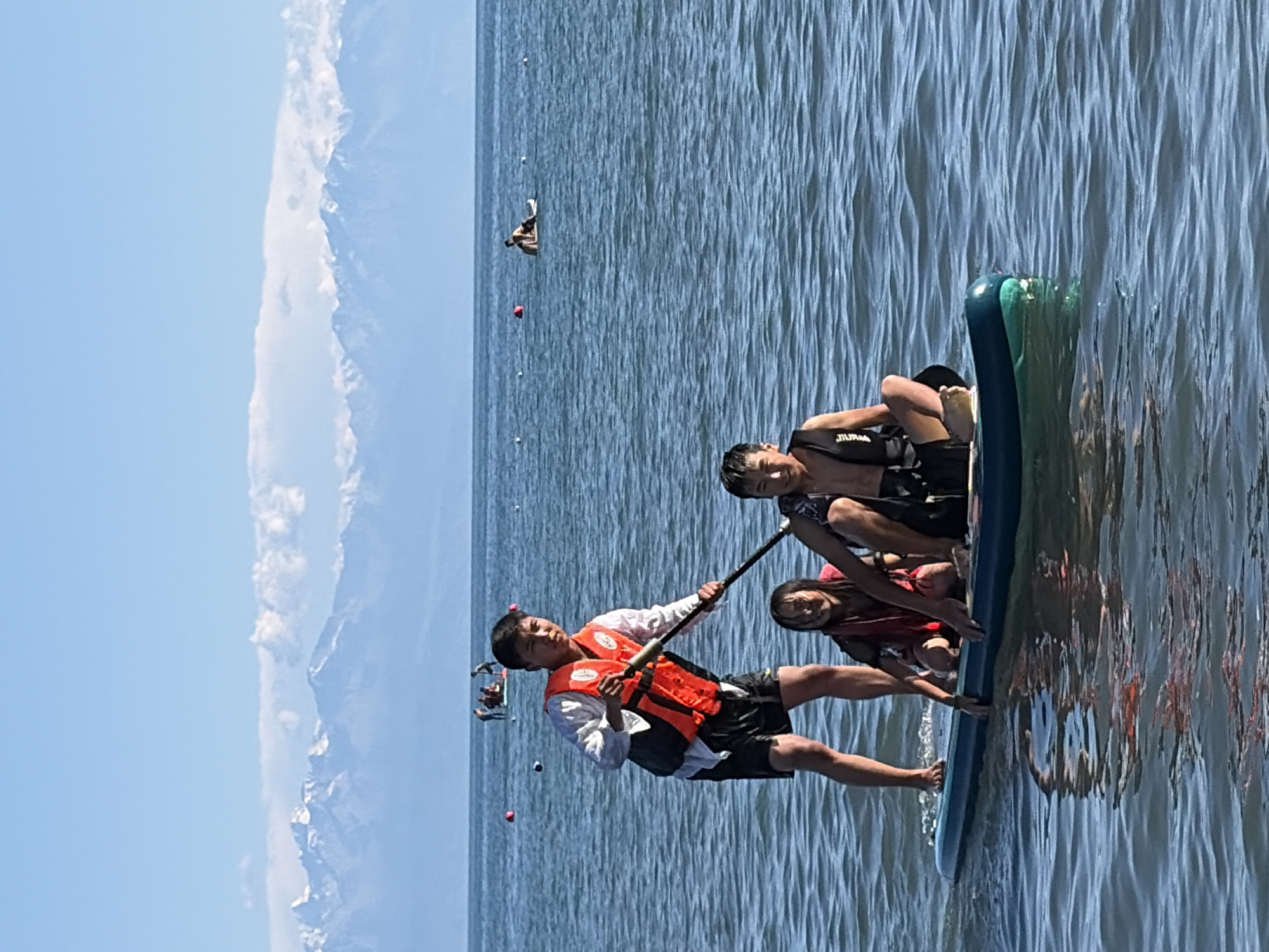 In the foreground, a young Asian male stands on a paddleboard, wearing an orange life vest and black shorts, holding a paddle. He is looking towards the camera. To his right and seated on the paddleboard are a young Asian female and a young Asian male, both wearing life vests. The paddleboard is floating on a body of water, with gentle ripples. In the background, several other people are visible on paddleboards in the water, with buoys marking an area. Beyond the water, a range of snow-capped mountains are visible under a clear blue sky with some clouds. The overall setting appears to be a recreational area on a lake or calm sea.