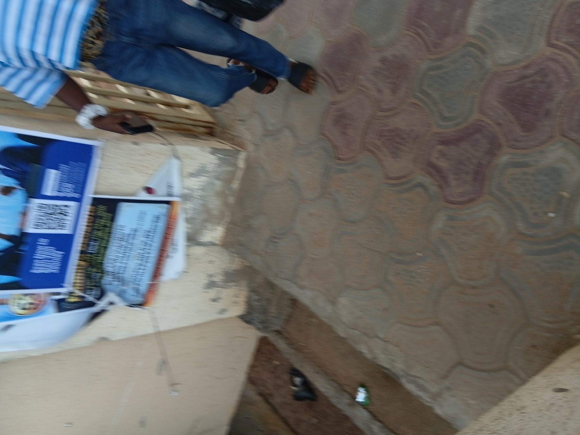 The image shows a person from the waist down, wearing blue jeans, a blue and white striped shirt, and sandals, walking on a patterned concrete sidewalk. The person is holding a dark-colored object, possibly a phone, in their right hand. A beige wall with peeling paint and posters is visible to the left. Some debris, including a black plastic bag, is on the ground near the wall. The background suggests an outdoor urban environment. The image is blurry due to motion. This scene is consistent with an everyday street scene in Jalingo, Nigeria.