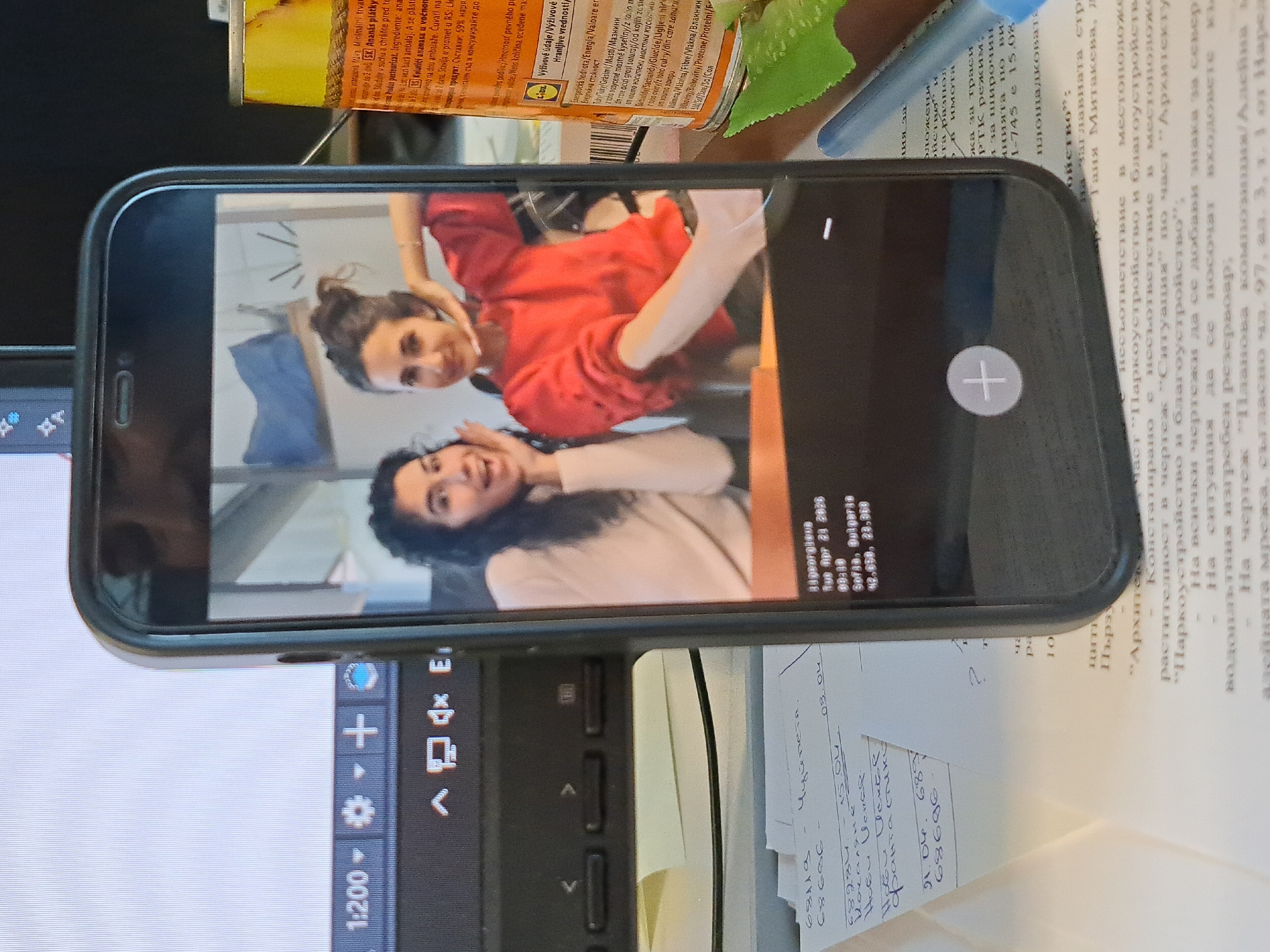 The media file displays a black smartphone lying on a desk, prominently showcasing a vertical image of two smiling young women with dark hair. The woman positioned higher in the photo wears a red top and has her hands playfully near her face, while the woman below her wears a light-colored top, striking a similar cheerful pose. The background behind them appears to be an indoor setting with light-colored walls.
On the smartphone's screen, an overlay of text indicates metadata for the displayed image: "Higeorgieva," taken on "Tue Apr 23 2029" at "09:30" in "Sofia, Bulgaria," with geographical coordinates "42.658, 23.98." A large white plus icon is also visible on the phone screen, likely an interface element for a photo gallery or editing app.
The phone is situated amidst a cluttered workspace. To its left, a segment of a laptop screen is visible, displaying interface elements including a "1:200" scale indicator. To the right, several documents with text primarily in Cyrillic (Bulgarian) are spread across the desk. These documents contain phrases related to "архитектура" (architecture) and "паркоустройство и благоустройство" (landscaping and urban planning), mentioning "Таня Митакева." Handwritten notes with dates such as "15.04" and "21.04" are also present on the papers. A yellow-labeled can, identified as "Ananas plátky" (pineapple slices) from LIDL with nutritional information, is partially visible at the top of the frame. A blue pen and a green, leaf-like object complete the visible items on the desk. The scene is lit by ambient indoor light, suggesting an office or home study environment.