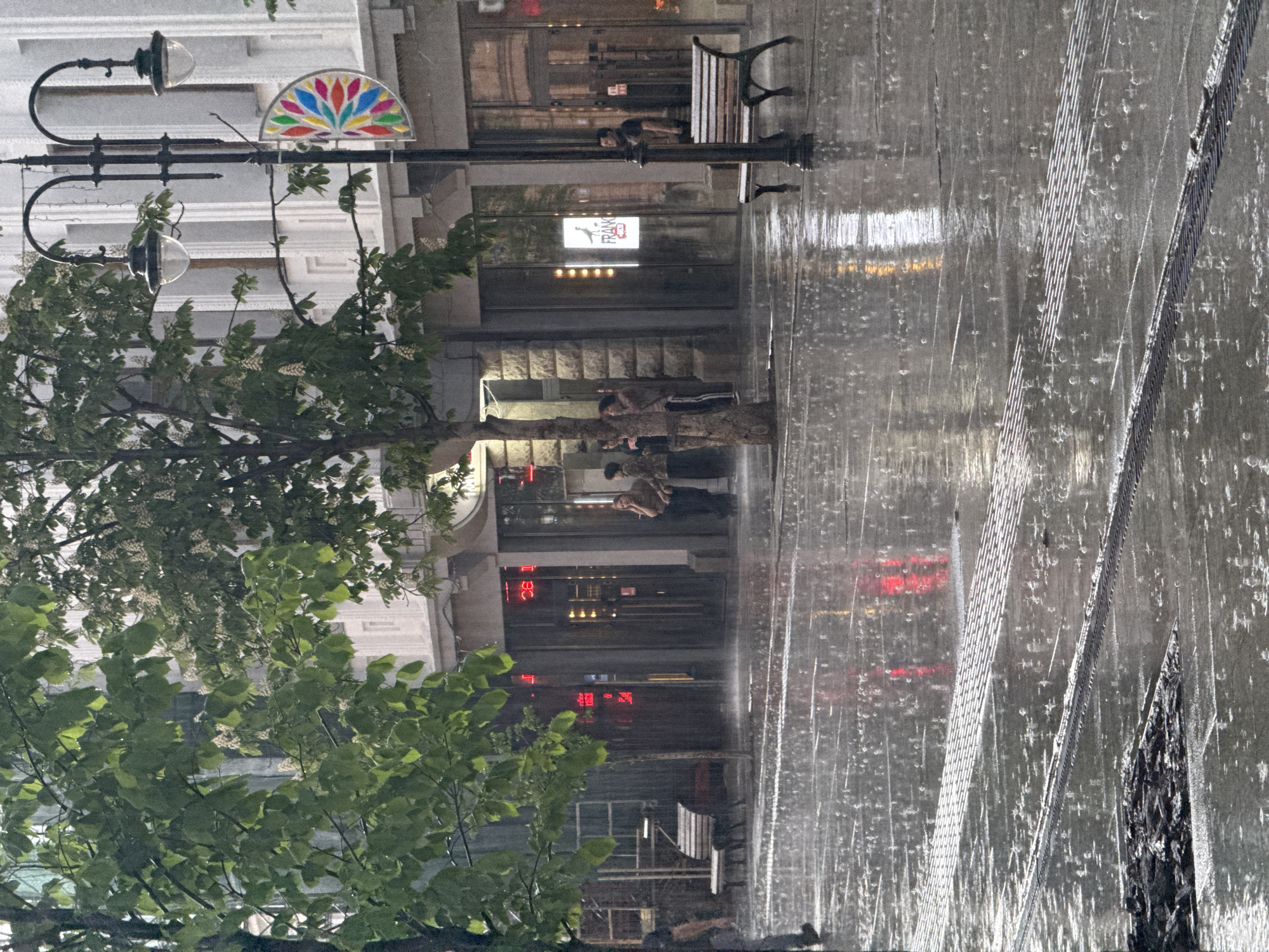 The image shows a rainy street scene in Saratov, Russia. Rain streaks obscure the view, but visible are buildings with signs, a tree with green leaves on the left, street lamps, and metal grating on the pavement. Several people are standing or sitting outside storefronts. A bench sits next to one of the buildings. Reflections of red and white lights are seen on the wet pavement.