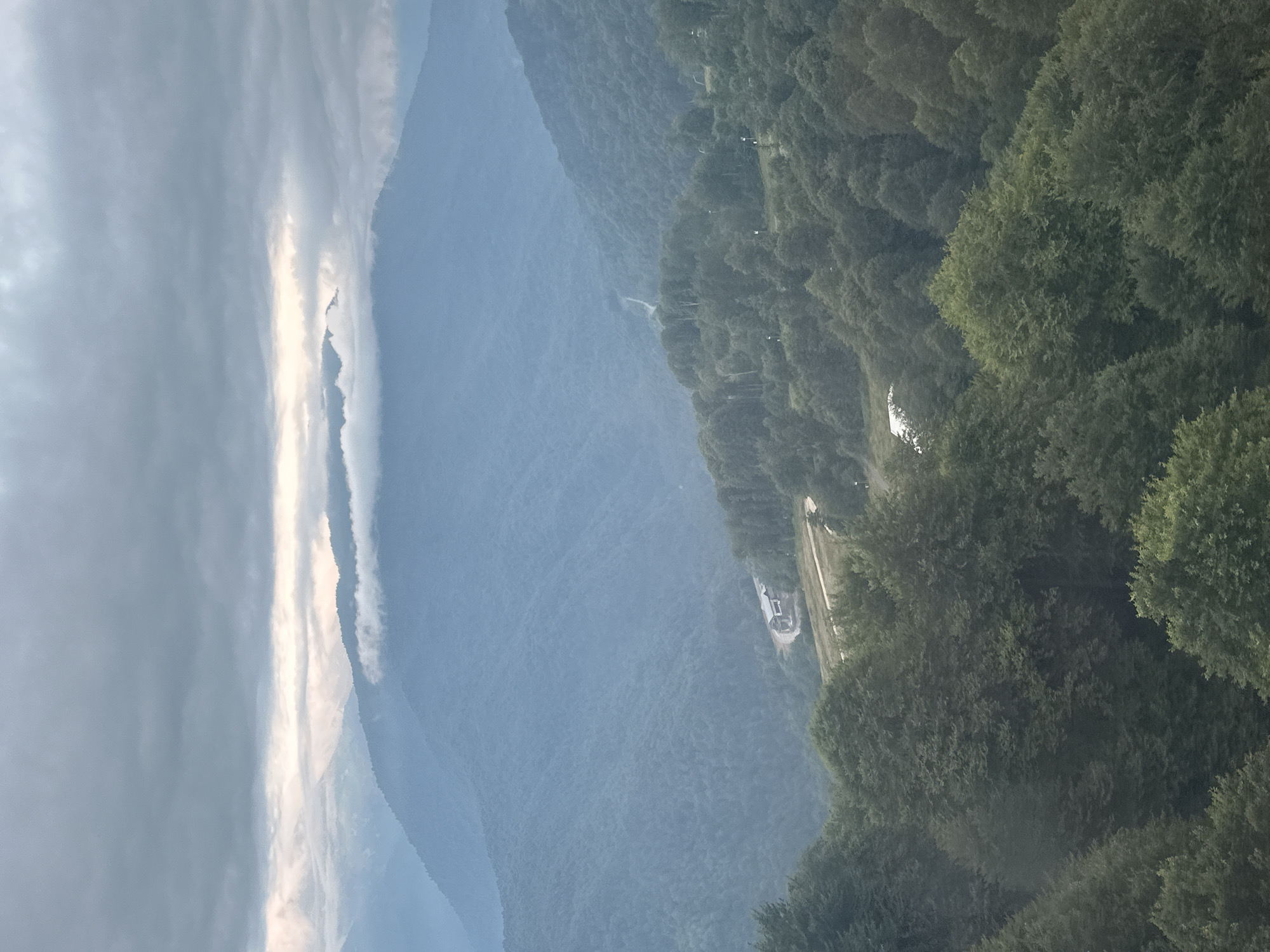 This is an aerial photograph of the mountainous landscape near Estosadok, Russia, under a cloudy sky. The image depicts several layers of misty, blue-hued mountain ranges receding into the distance. The foreground is dominated by dense, dark green forests, with a winding dirt road visible leading to a white building nestled on a slope in the mid-ground. The sky is overcast with dramatic, heavy clouds, with a sliver of brighter sky visible between the cloud layers and the distant mountains. There are no people visible in the image.