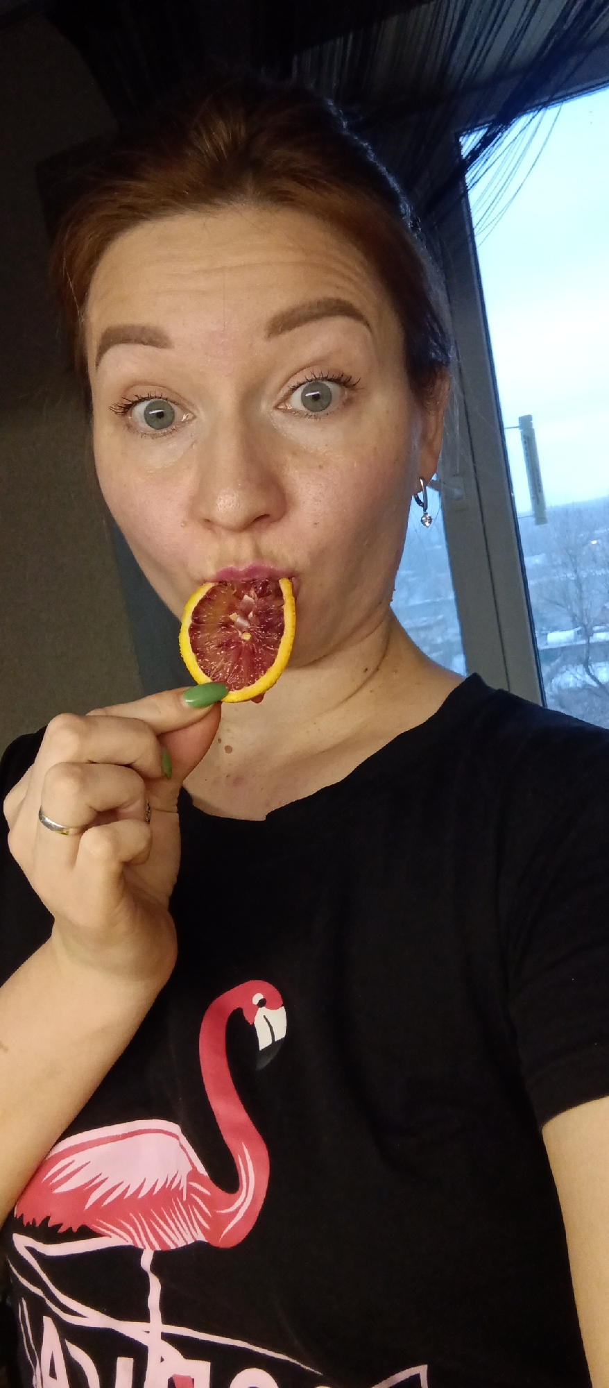 A selfie shows a Caucasian woman with reddish hair and blue eyes holding a slice of blood orange in front of her mouth. She has on an earring and a black t-shirt with a flamingo design. In the background, there is a window with a view of a cityscape with snow on the ground. The city is Unknown, Unknown.