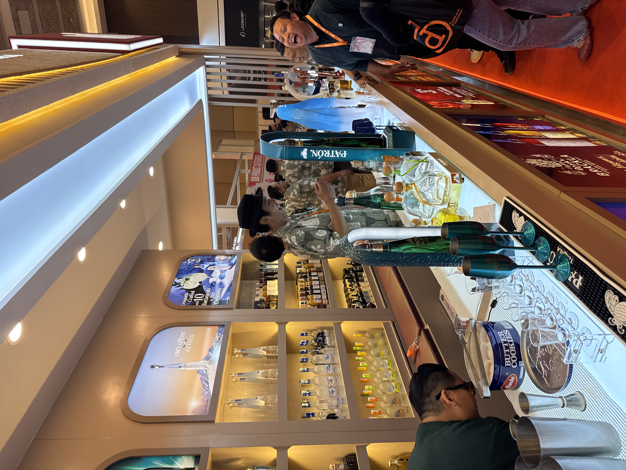 This image captures an indoor scene, likely a commercial event or a high-end bar setup, featuring several individuals and extensive displays of alcoholic beverages in Kuta Selatan, Indonesia.
In the foreground, a man wearing a dark green shirt and glasses is visible from behind, seemingly tending to a counter. The counter features bar equipment including stainless steel shakers, a jigger, and various clear glasses. A blue tin labeled "BUTTER COOKIES" by Kjeldsens sits on the counter. Behind him, another man in a patterned shirt stands near a display of clear and amber-colored bottled spirits.
Further into the scene, a man in a dark t-shirt with a lanyard around his neck is laughing heartily, looking towards the camera. He stands beside a counter lined with numerous bottles and illuminated panels displaying text and graphics. The flooring in this area is a vibrant red carpet.
The background reveals a sophisticated display area with shelves filled with various bottles of alcohol. Illuminated display panels are integrated into the walls, showcasing brands like "GREY GOOSE ALTIUS" and an ad stating "Uniquely Crafted with 10 Botanicals to Stir Creativity." A prominent display for "PATRÓN." tequila is visible, featuring a large, ornate bottle and branded items. Other visible brands include "JOSPER" on equipment and "PATRON" and "EL CIELO" on a counter mat. The overall lighting is artificial, with strip lighting illuminating the displays and spotlights in the ceiling, creating a bright and inviting atmosphere. The scene suggests a lively product showcase or tasting event.