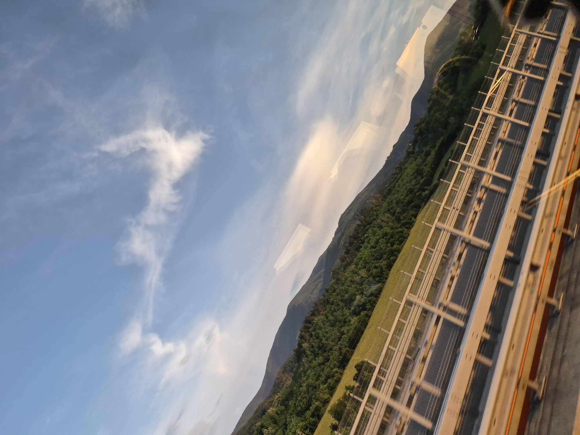 The image captures a scenic view of rolling green hills and mountains under a bright blue sky with scattered wispy clouds. In the foreground, a highway guardrail is prominently visible, suggesting the photo was taken from a moving vehicle on a road. The sunlight creates highlights and reflections on what appears to be a window, adding a slightly distorted perspective to the scene. The lush green vegetation covering the hills indicates a natural, possibly rural or mountainous environment. The location is identified as the city of Razliv, Bulgaria.