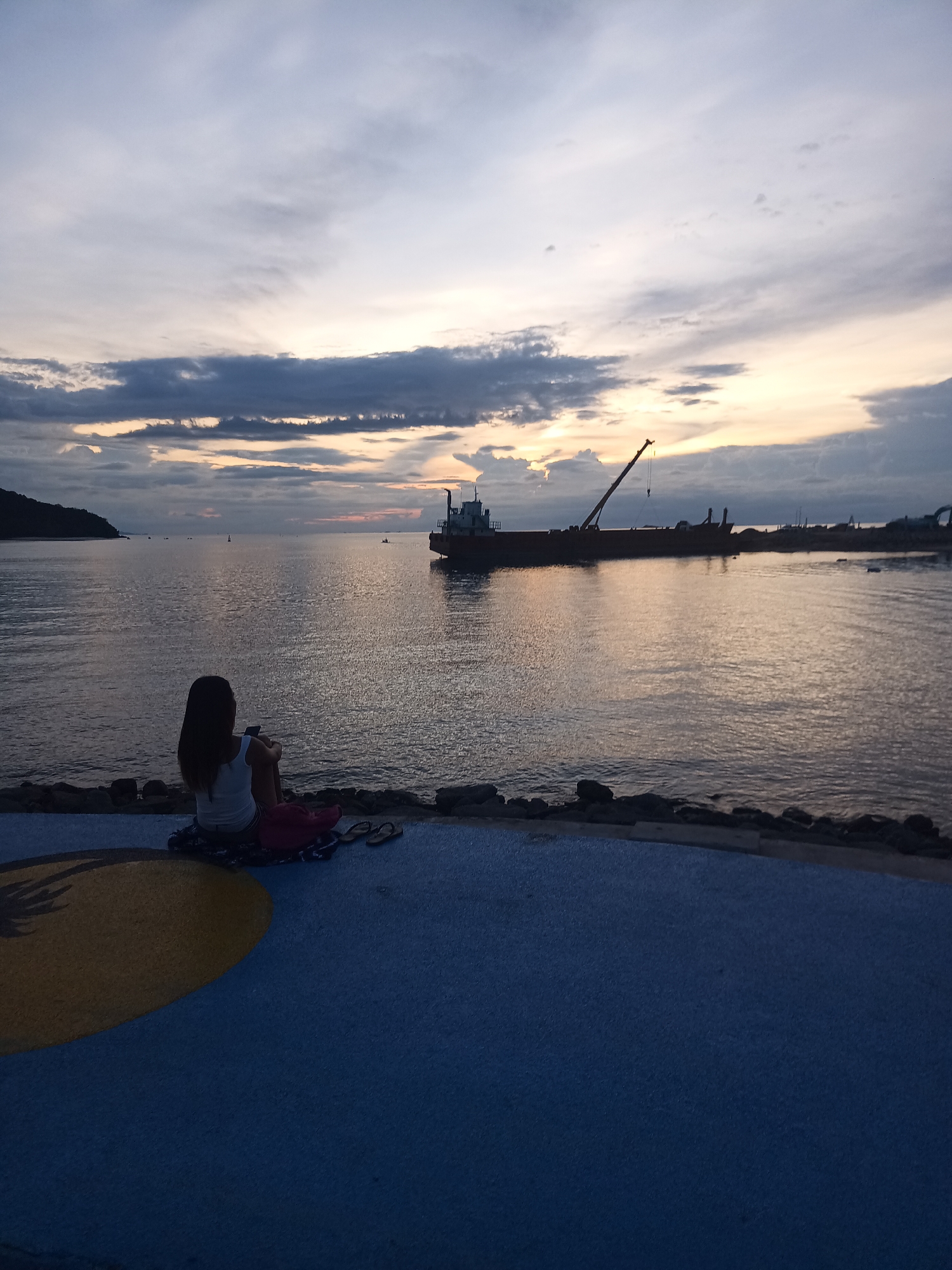 A woman is sitting on a blue and yellow painted surface, facing away from the camera, towards a calm body of water. She is wearing a white tank top and dark shorts. A pink blanket and a pair of flip-flops are on the ground next to her. In the distance, a large ship with a crane is visible on the water, with the sun setting behind dark clouds in the sky. The scene appears to be a waterfront public space in Koh Phangan, Thailand.