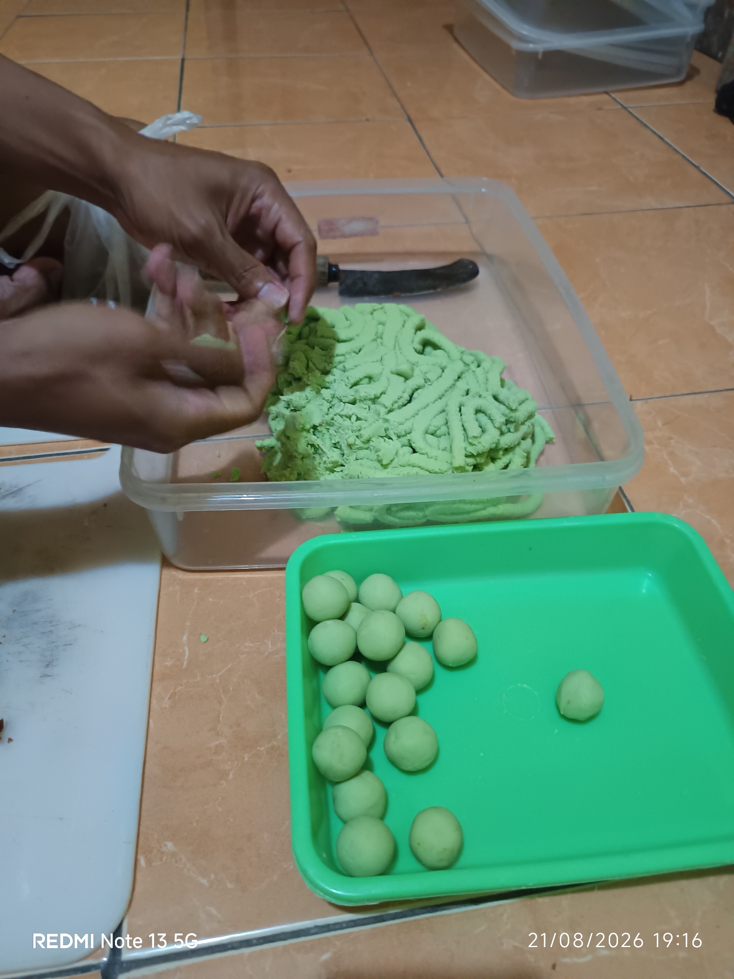 In Talaga Sari, Indonesia, an individual is shown preparing a green, dough-like substance. The hands are actively working with a portion of the green material, suggesting it is being shaped or processed. The substance is contained within a clear plastic tub, and next to it is a knife. Below this, a bright green rectangular tray holds several small, round, green balls, indicating a stage of completion in the preparation process. The surrounding environment appears to be an indoor setting with tiled flooring.