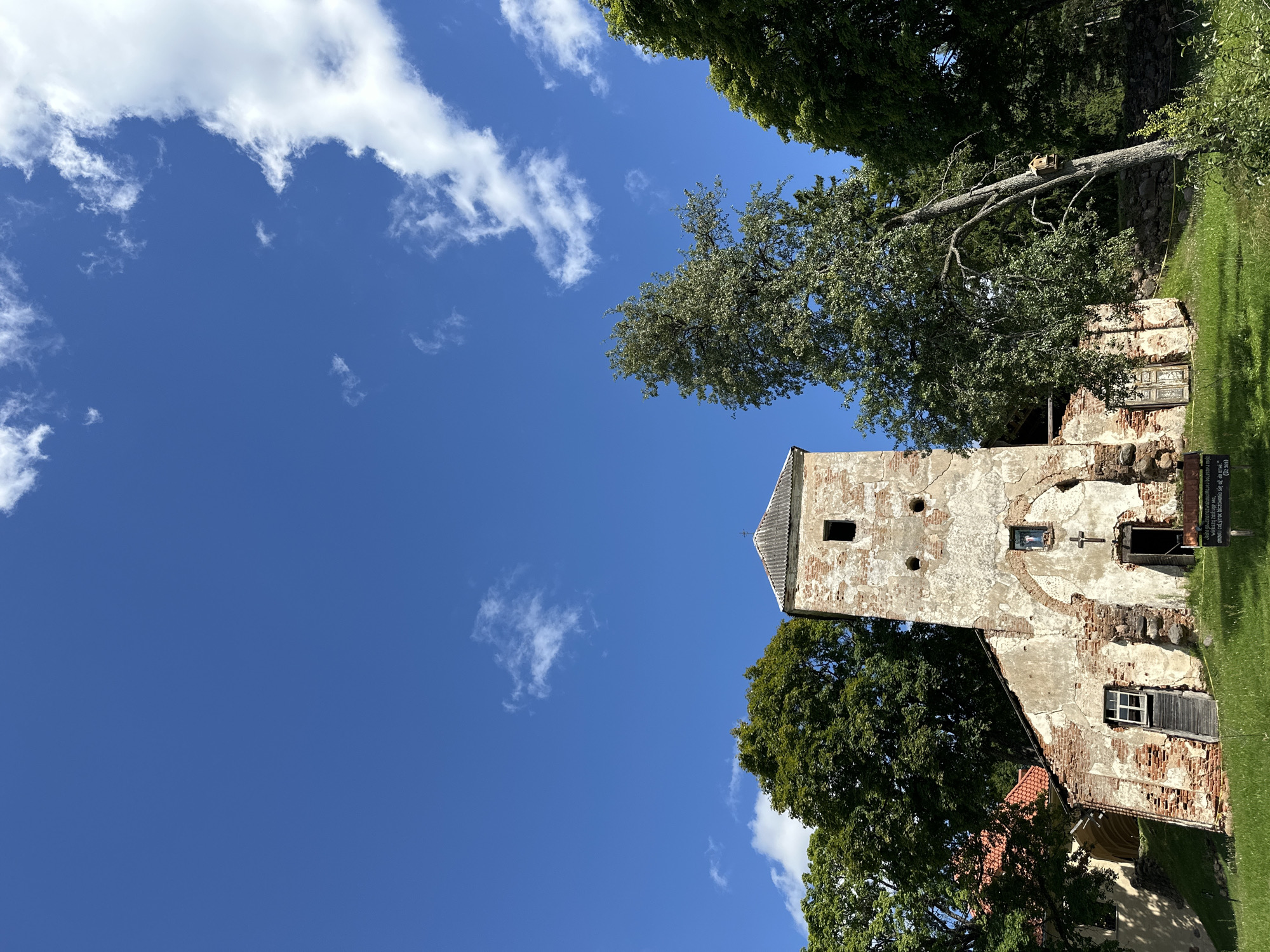 The image depicts the dilapidated ruins of a church tower in Norviliškės, Lithuania. The brick tower, largely unplastered and showing signs of decay, stands tall against a bright blue sky dotted with white clouds. A simple, grey roof caps the tower. The facade features a small, dark rectangular window near the top and two circular openings below it. Further down, a smaller rectangular window is positioned above a crucifix. The main entrance at the base of the tower is open, revealing a dark interior. Adjacent to the tower, an archway leads to a more intact section of the building, partially visible with a red roof. A wooden staircase leads up to a small window on the side of the tower. The ground in front of the structure is covered in green grass, and several trees with lush green foliage surround the ruins. To the right of the tower, a slender tree leans precariously, with a birdhouse attached to its trunk. In the foreground, a yellow caution tape is visible, and a sign with text is placed on the grass. The overall scene conveys a sense of historical neglect and natural overgrowth.