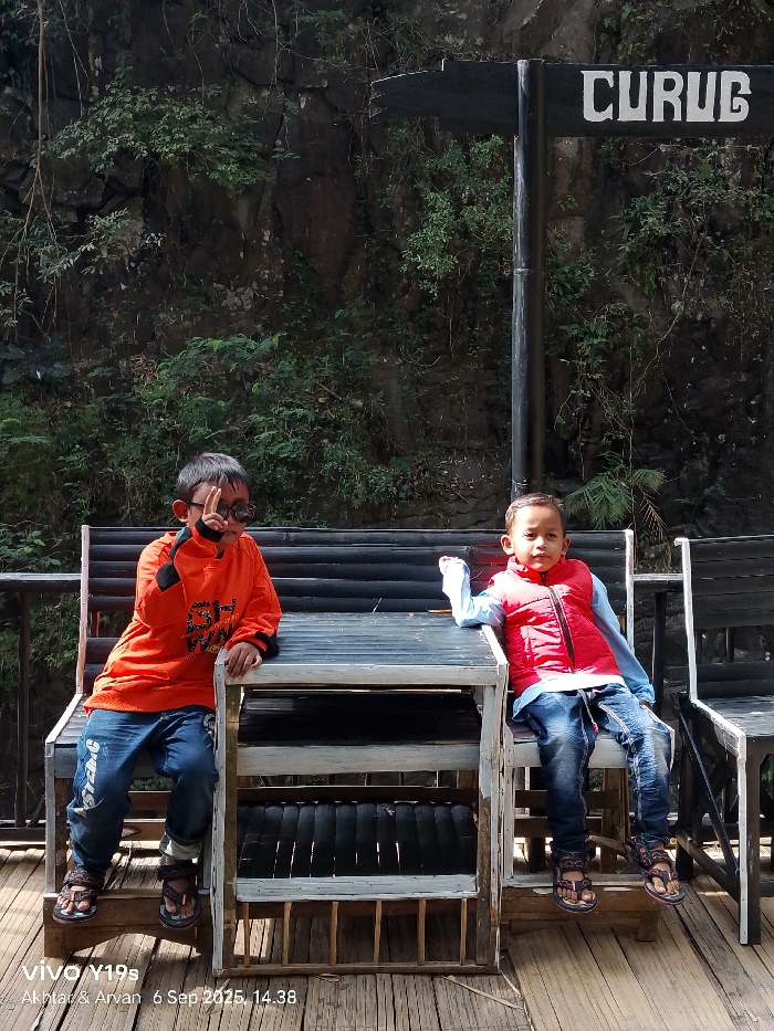 Two young boys are seated on rustic black and white wooden benches around a central wooden table. The scene is located in Sukadana, Indonesia, near a waterfall, as indicated by a black signpost bearing the white text "CURUG" against a background of a dense, rocky, vegetated cliff face. The boy on the left wears an orange long-sleeved shirt, blue jeans, and dark sunglasses, making a peace sign with his right hand. The boy on the right wears a light blue long-sleeved shirt, a red puffer vest, and blue jeans, looking directly forward. Both boys are wearing sandals. The ground beneath them consists of a light-colored slatted wooden platform. The image was captured on September 6, 2025, at 14:38.