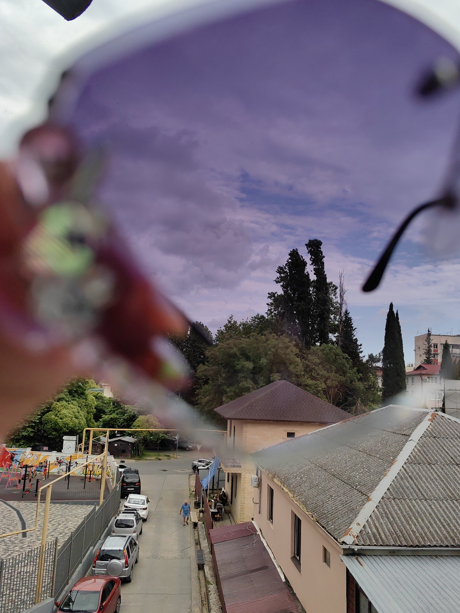 The image, taken from a high vantage point, depicts a street scene in Sochi, Russia. The foreground is dominated by a blurred view through the lens of a pair of sunglasses, showing a purple-tinted sky with clouds. Below, a narrow street is lined with parked cars. A playground area with red and yellow structures is visible to the left, bordered by a metal fence. Buildings with tiled roofs are present on either side of the street, with some taller trees and cypress-like evergreens in the background. A single person in a blue shirt and shorts is walking down the street. The overall impression is a residential area with a playground, set against a backdrop of lush greenery and a cloudy sky.