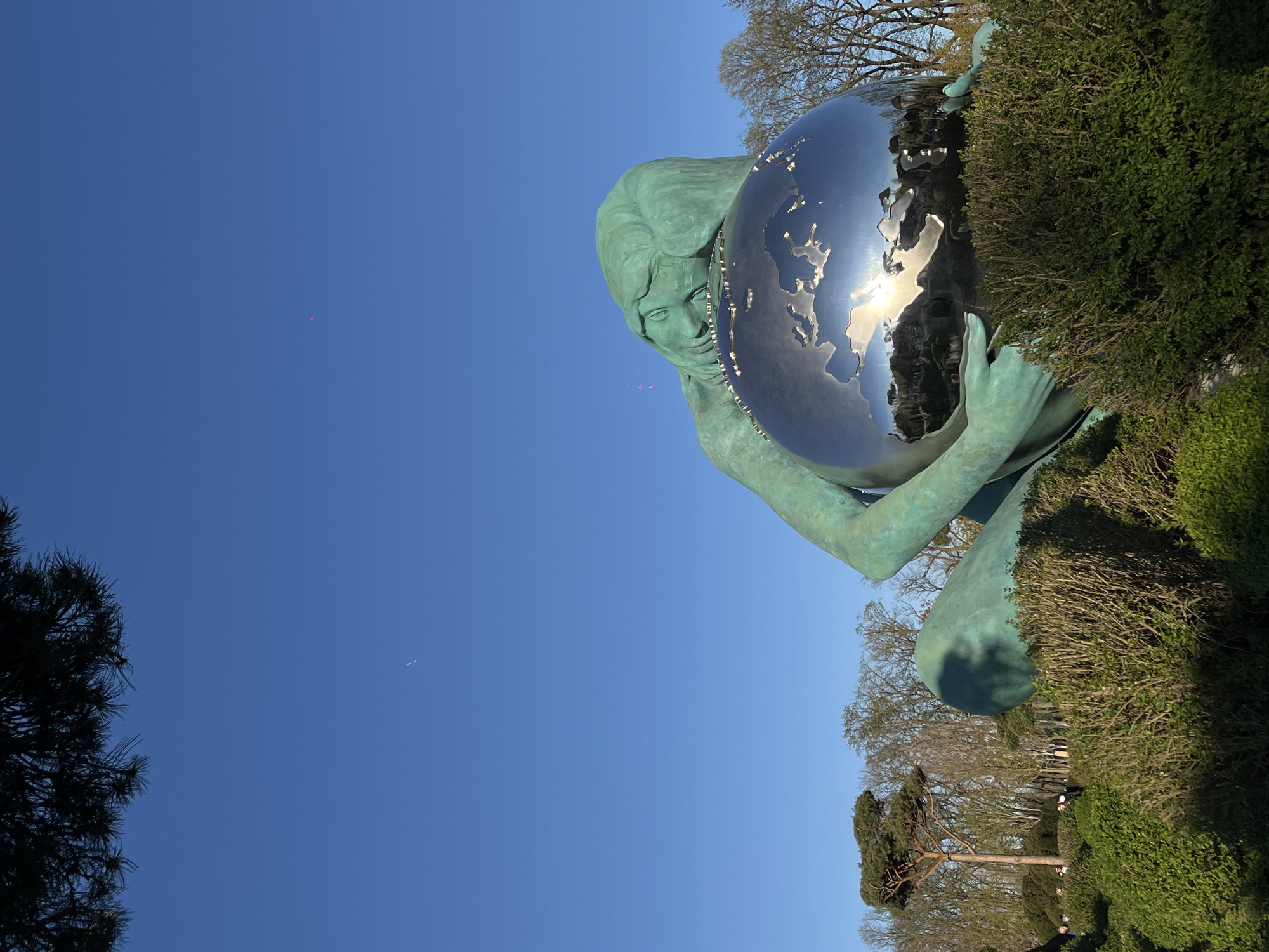 Here is a detailed description of the image:
The image shows a sculpture in the city of Krasnodar, Russia. The sculpture depicts a woman holding a large, metallic globe. The woman appears to be made of a green, weathered material, possibly bronze or copper that has oxidized. The globe is highly reflective, mirroring the surrounding environment, including the sky and possibly the sun. The sculpture is situated in a park or garden setting, with manicured bushes and trees visible in the background. The sky is a clear, bright blue. Several small, pink objects are floating in the air, perhaps balloons or confetti. The scene is photographed from a low angle, looking up at the sculpture.