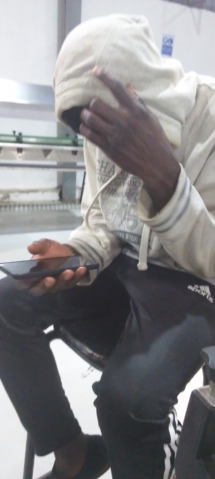 A person wearing a light-colored hoodie, pulled over their head to obscure their face, sits on a chair in an indoor setting. They are holding a black smartphone in their left hand. The person is wearing dark-colored sweatpants with white stripes down the side of the legs, and black footwear. In the background, a green metal structure is visible, along with a white wall. A partially obscured sign that says "06" is also visible on the white wall. The specific location in Ikorodu, Nigeria, is not discernible from the image.