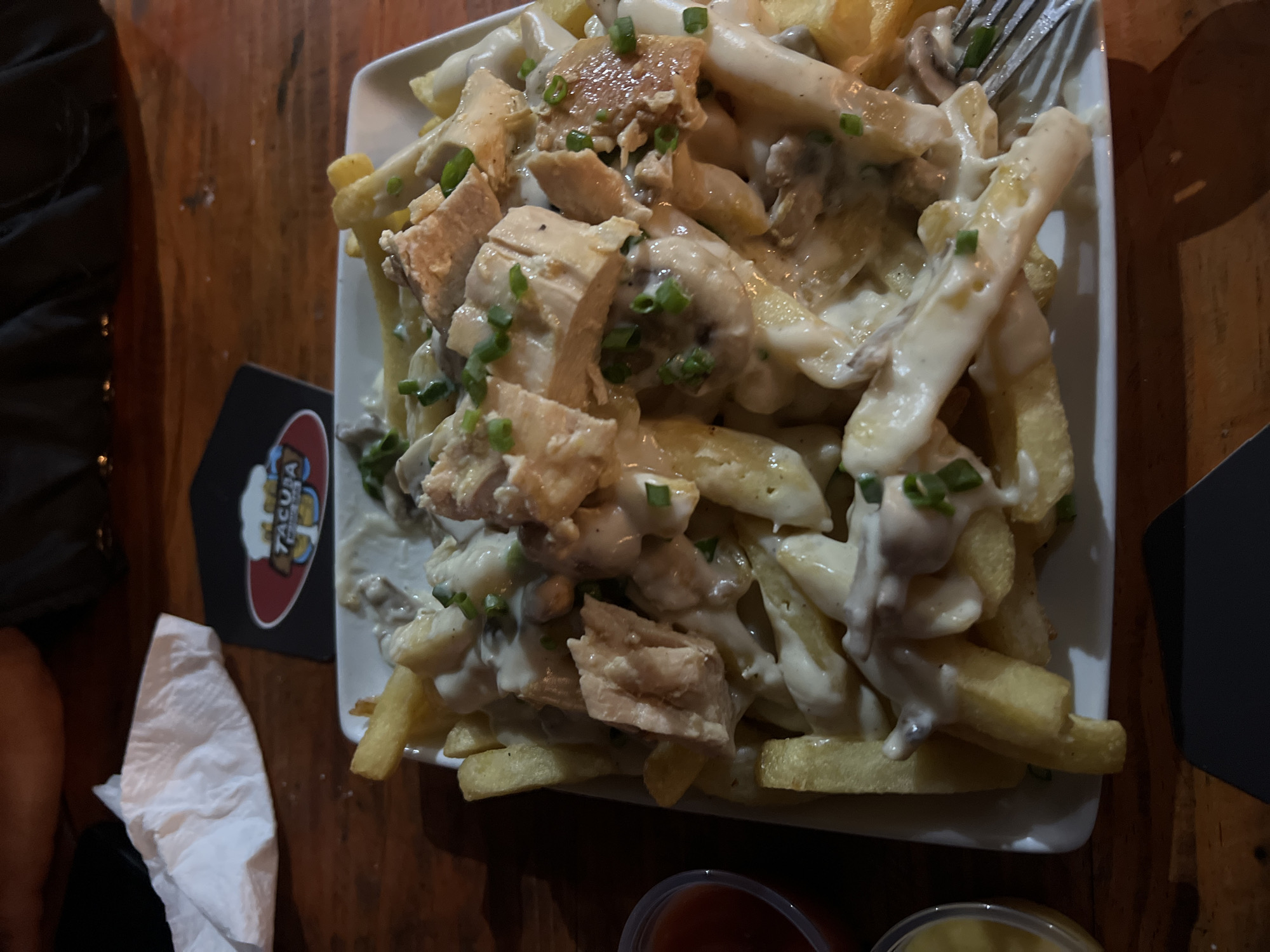The image shows a plate of fries topped with chicken, mushrooms, and a creamy sauce, sprinkled with chopped green onions. A fork rests on top of the food. The plate is placed on a wooden table, next to a coaster with the logo "TACURA" visible. A crumpled white napkin and a black object are on the left side of the plate, and two small containers of condiments are visible at the bottom. Based on the "TACURA" coaster, the location is likely in Arica, Chile, as "Tacura" is a restaurant and bar in that city. There are no people visible in the frame besides a partial view of a person's leg/arm.