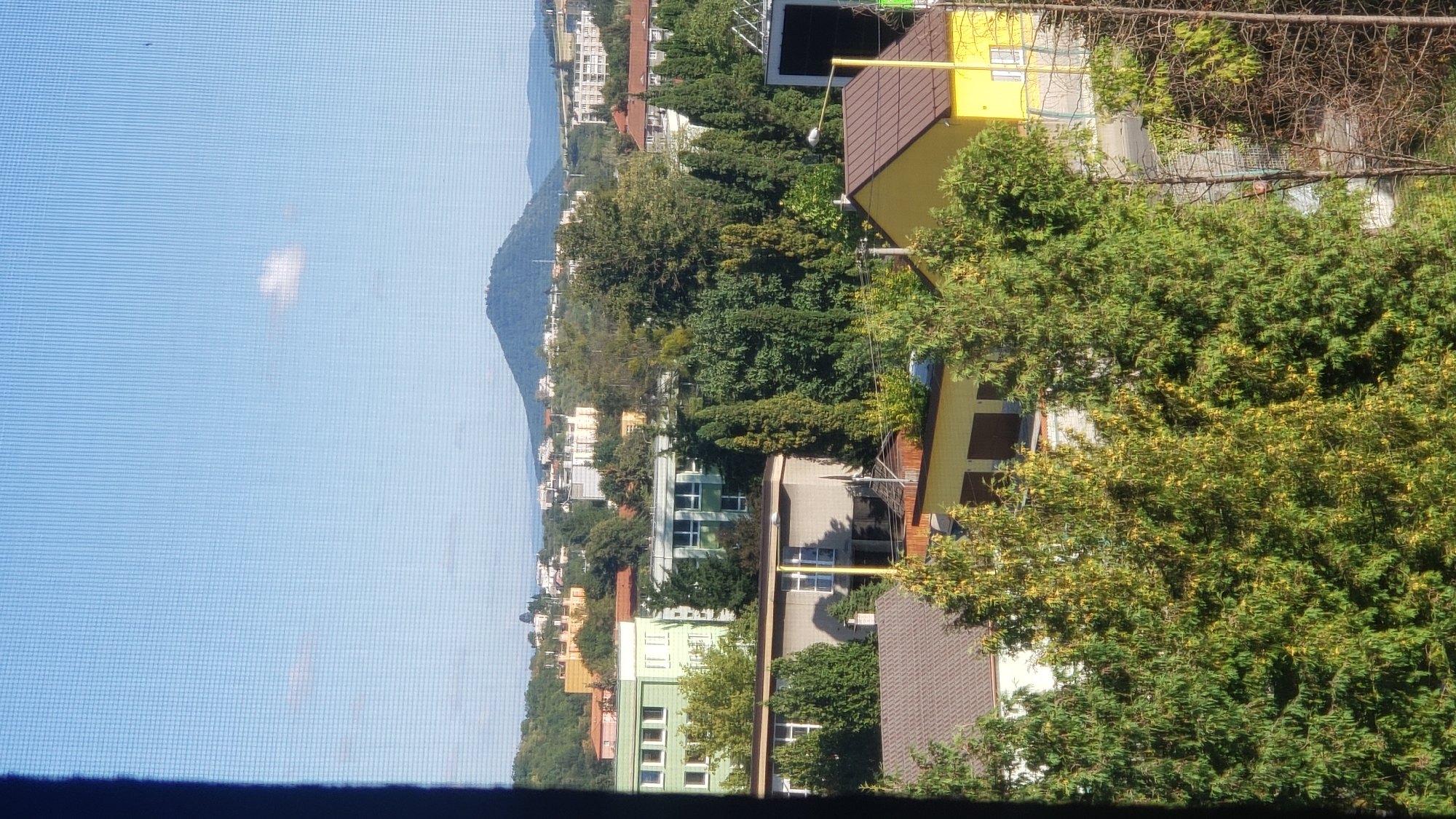 The image captures a view of the city of Prešov, Slovakia, as seen through a window with a visible screen mesh. In the foreground, lush green trees dominate, with some showing hints of yellow foliage, suggesting it is late summer or early autumn. Behind the trees, a collection of buildings is visible, including a light brown, multi-story structure with visible windows and a dark brown roof. To its right, a bright yellow building with a brown roof features windows. Further back, several apartment buildings are scattered among more trees, with colors ranging from green to various shades of orange and white. In the distance, a prominent, rounded, tree-covered hill or mountain rises against a clear blue sky with a single, small white cloud. Other smaller hills are visible in the far background. The overall scene depicts a semi-urban environment with a blend of residential buildings and natural landscape.