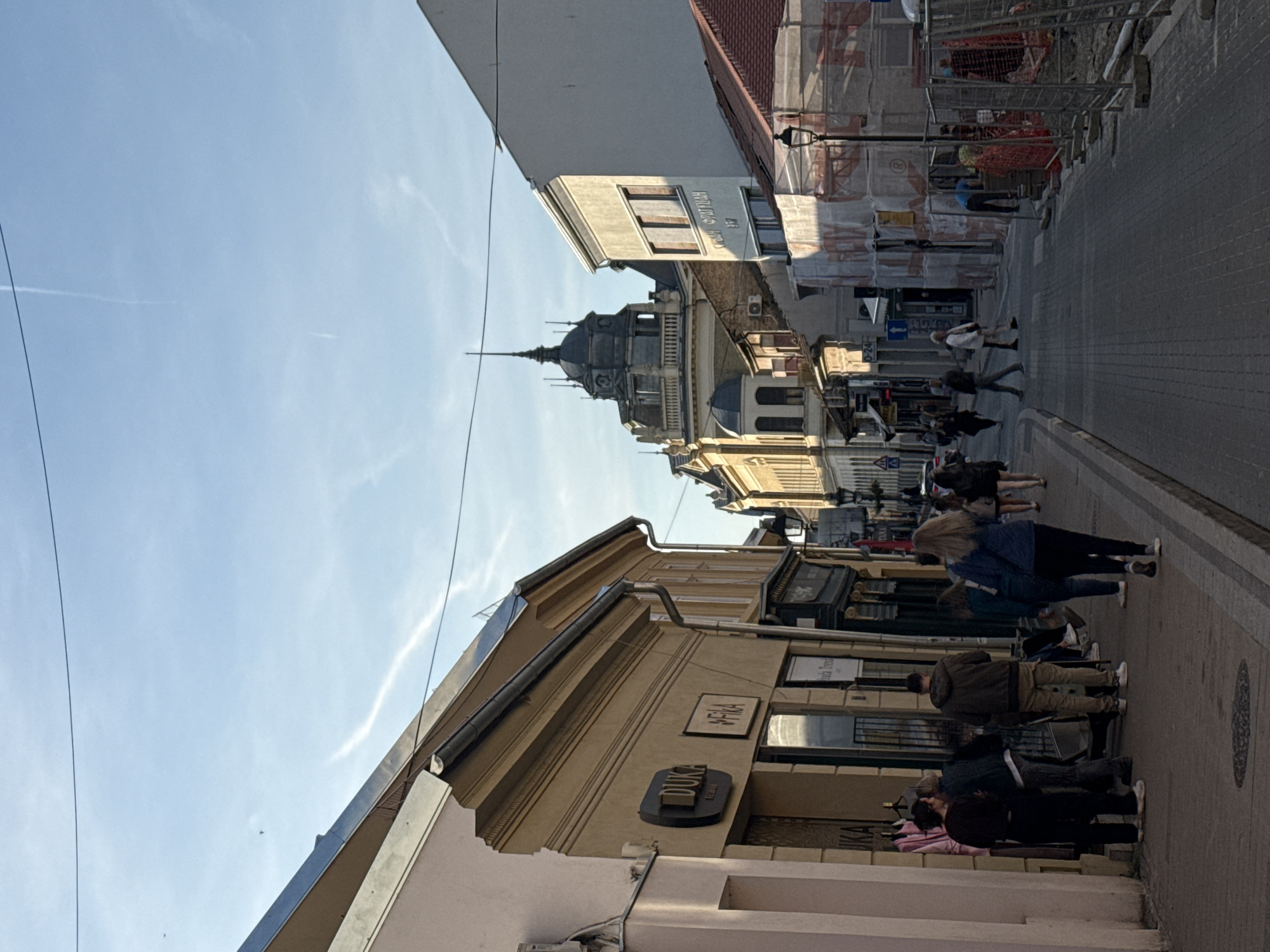 Here's a description of the image:
This is an eye-level outdoor shot on a pedestrian street in an urban setting. The view is looking down the street between buildings on either side. On the left, a tan-colored building with a sign that says "Duka" is visible. On the right, taller buildings are present, some under construction with scaffolding. People are walking along the street on the right side. In the distance, an ornate building with a tower stands out. The sky is visible above the buildings, showing a clear blue with some wispy clouds and vapor trails. The pavement appears to be made of brick. The location is in the city of Unknown, Unknown.