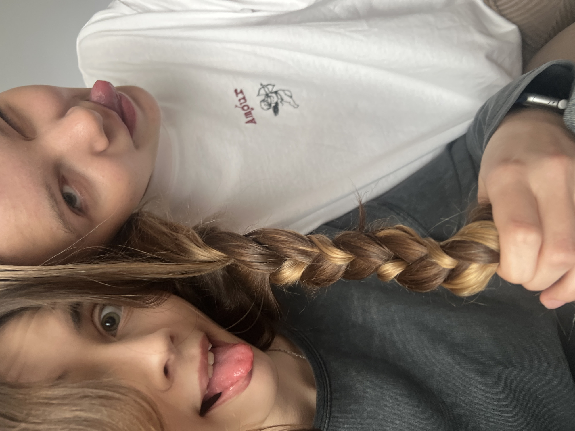 Two individuals are positioned closely, with the face of one partially obscured by the other's hair, which is styled in a blonde and brunette braid held by a hand. Both individuals are sticking out their tongues. One person is wearing a white shirt with a logo, and the other is wearing a grey top. The location is not specified as Votkinsk, Russia, nor is there enough information to determine a city.