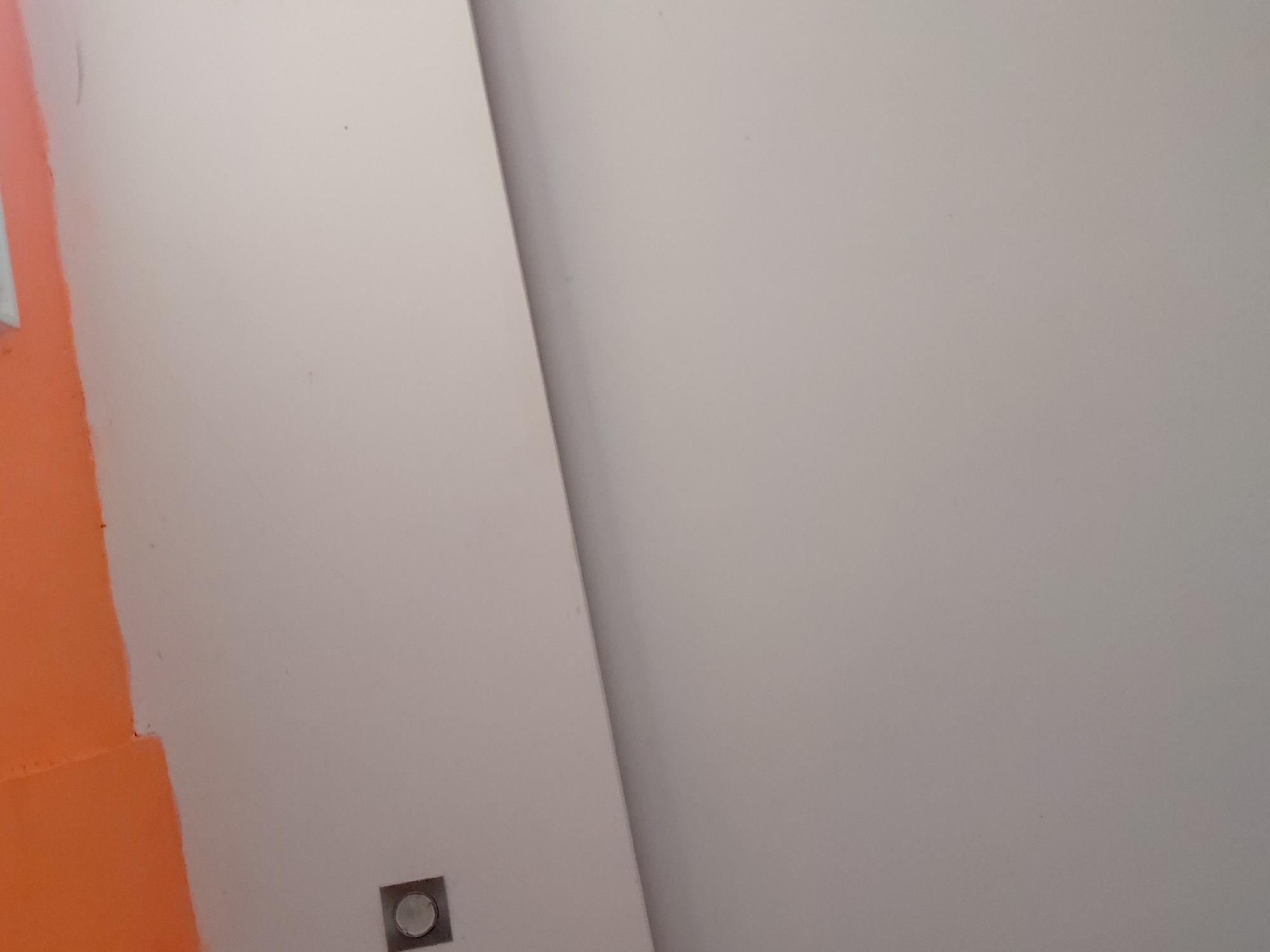 The image displays an indoor scene, characterized by a minimalist architectural aesthetic. No people are visible. On the left side, a vibrant orange wall is partially depicted, contrasting with a larger, off-white or light gray wall that occupies most of the frame. A distinct diagonal line runs across the image, marking the intersection or separation of these two wall surfaces. Near the bottom left corner of the off-white wall, close to the orange section, a small, square-shaped fixture with a circular, metallic-looking center is flush-mounted. This object appears to be a light switch, an electrical outlet, or a small flush light. The walls appear to have a smooth, painted finish. The lighting is even, suggesting ambient indoor illumination. No specific activity or event is taking place, and the scene is static. There is no visible text. While the location context is provided as Santa Ana, Venezuela, there are no specific visual cues within the image itself to confirm this.