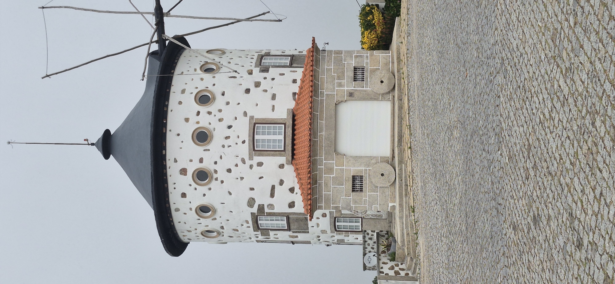 The image depicts a traditional stone and white-plastered windmill in Póvoa de Varzim, Portugal. The windmill has a conical, dark gray roof and its cylindrical body is adorned with round, small windows and irregularly placed stones. A portion of the windmill's blades are visible on the right side of the frame. At the base of the windmill, there is a section with a red-tiled roof supported by a stone structure. This section features a large, white roller door and two large millstones positioned on either side. The ground in front of the structure is paved with cobblestones. The sky is overcast.