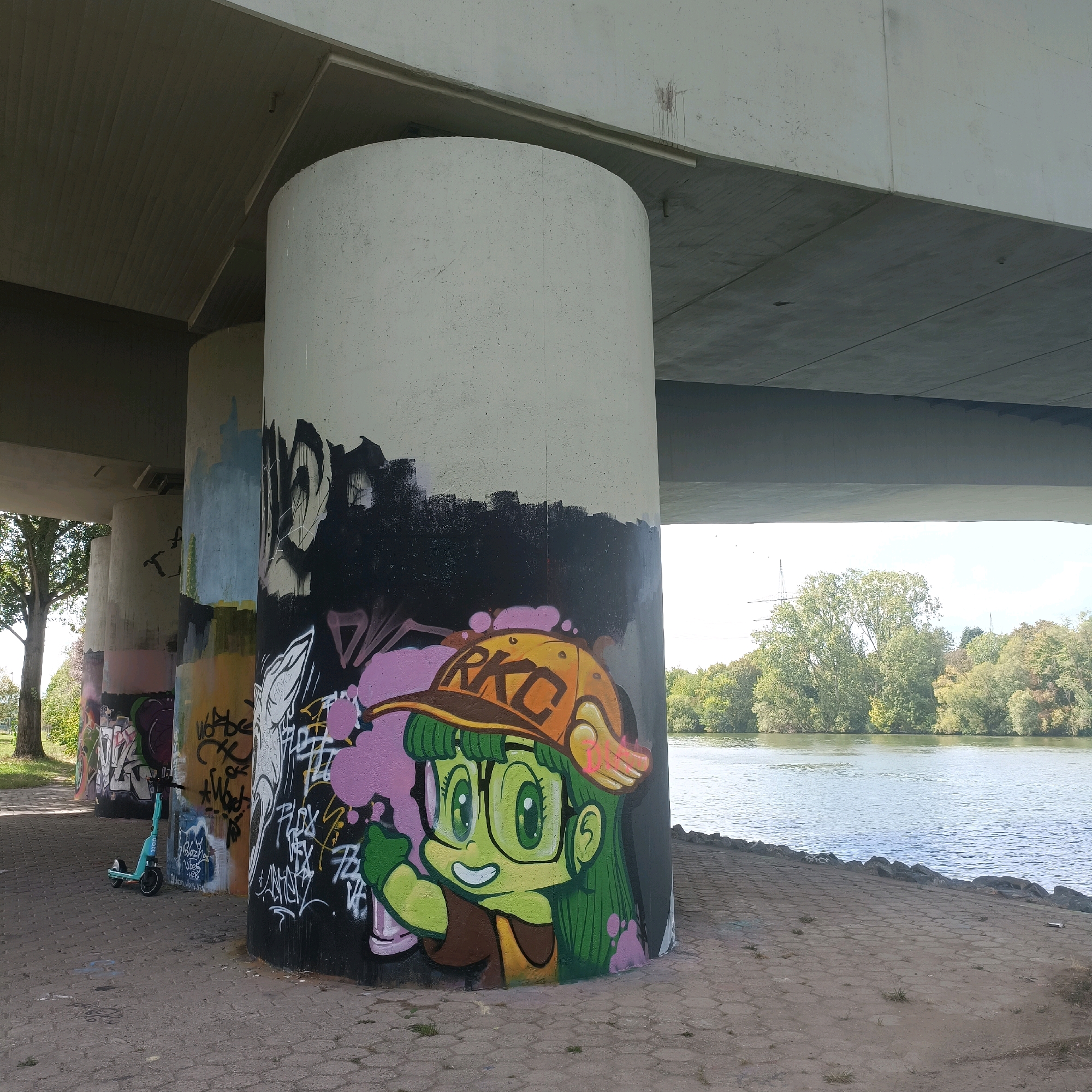 An outdoor photograph taken from a low angle under a bridge in Rüsselsheim am Main, Germany. The bridge is made of concrete with a textured ceiling. The scene is dominated by several large, cylindrical concrete pillars that support the bridge. These pillars are heavily adorned with graffiti. The most prominent graffiti is on the foreground pillar, a colorful depiction of a cartoon character with green hair, glasses, and an orange baseball cap with the letters "RKC" on it. The character is giving a thumbs-up. Other graffiti on the pillars includes abstract designs and lettering in various colors, such as black, white, pink, blue, and yellow.
To the left of the pillars, a light blue electric scooter is parked on a paved walkway. The walkway is made of light brown rectangular paving stones. Beyond the walkway and pillars, a body of water, likely the Main River, is visible. The water is calm with a slight ripple and reflects the sky and the lush green trees that line the opposite bank. The sky is bright and appears to be partly cloudy. The overall atmosphere of the scene is urban and somewhat edgy due to the graffiti, contrasted with the natural beauty of the river and trees.