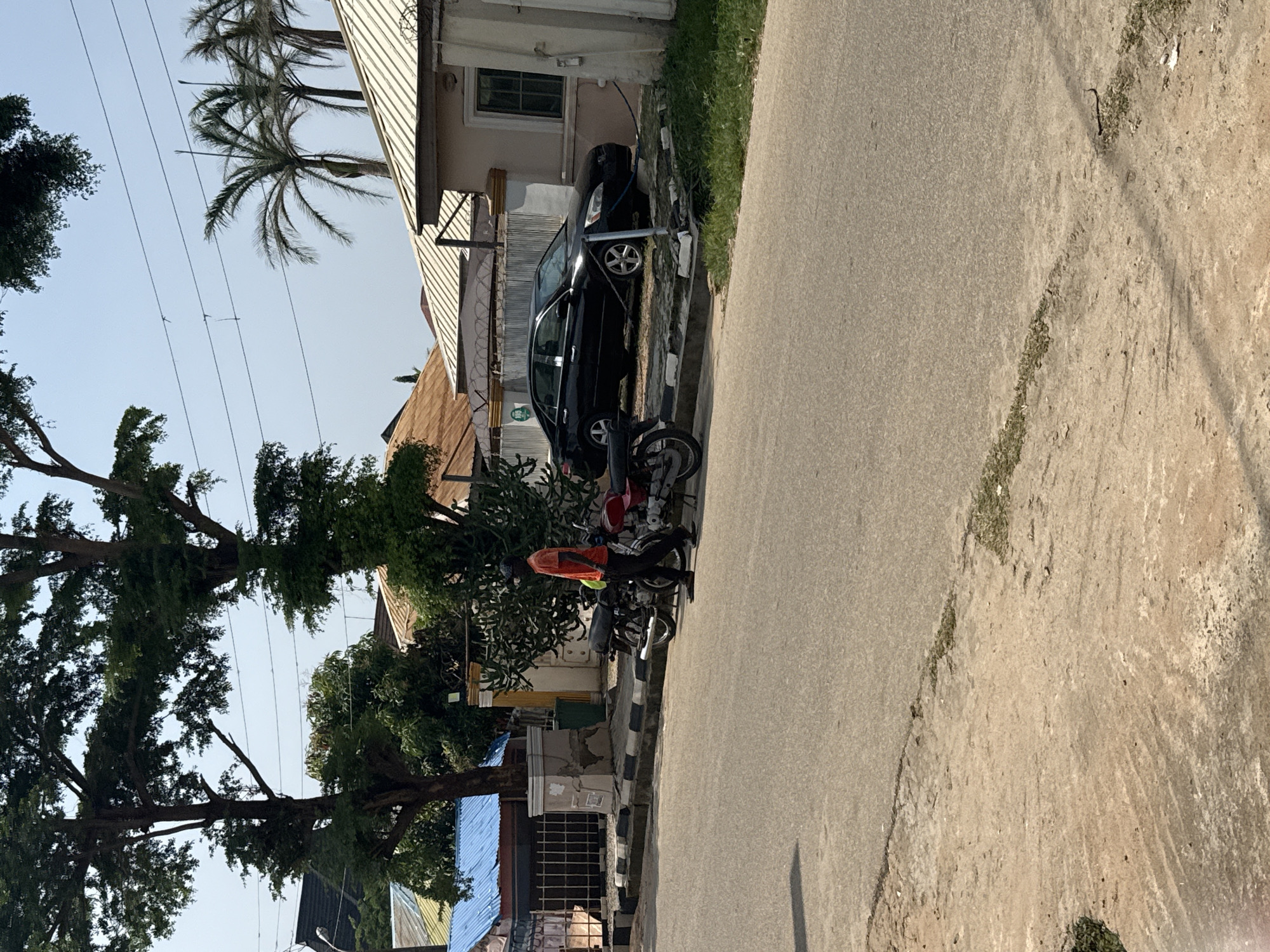 A man in an orange shirt and black pants walks past two parked motorcycles on a paved street in Abuja, Nigeria. A black sedan is parked in a driveway behind a metal fence. Tall trees with green foliage are visible in the background, along with palm trees and several buildings with light-colored roofs. The sky is clear and blue.