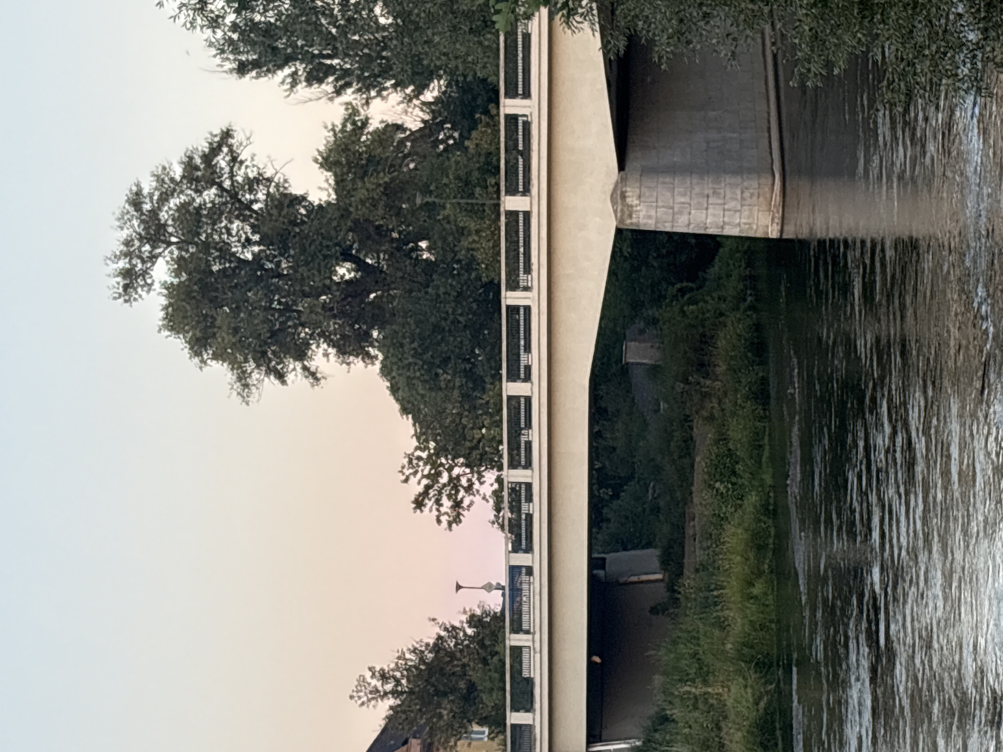 The image shows a wide, concrete bridge spanning a body of water. The bridge has a light-colored surface and a decorative railing with a black metal fence. The underside of the bridge is visible, revealing a dark opening. On the left side of the image, there is a building with a peaked roof and a window. A single street lamp is visible on the bridge. The area surrounding the water is covered in lush green vegetation, including tall grass and trees. The sky above is a soft gradient of pale pink and light blue, suggesting either sunrise or sunset. The water reflects the sky and the bridge, with ripples indicating gentle movement.