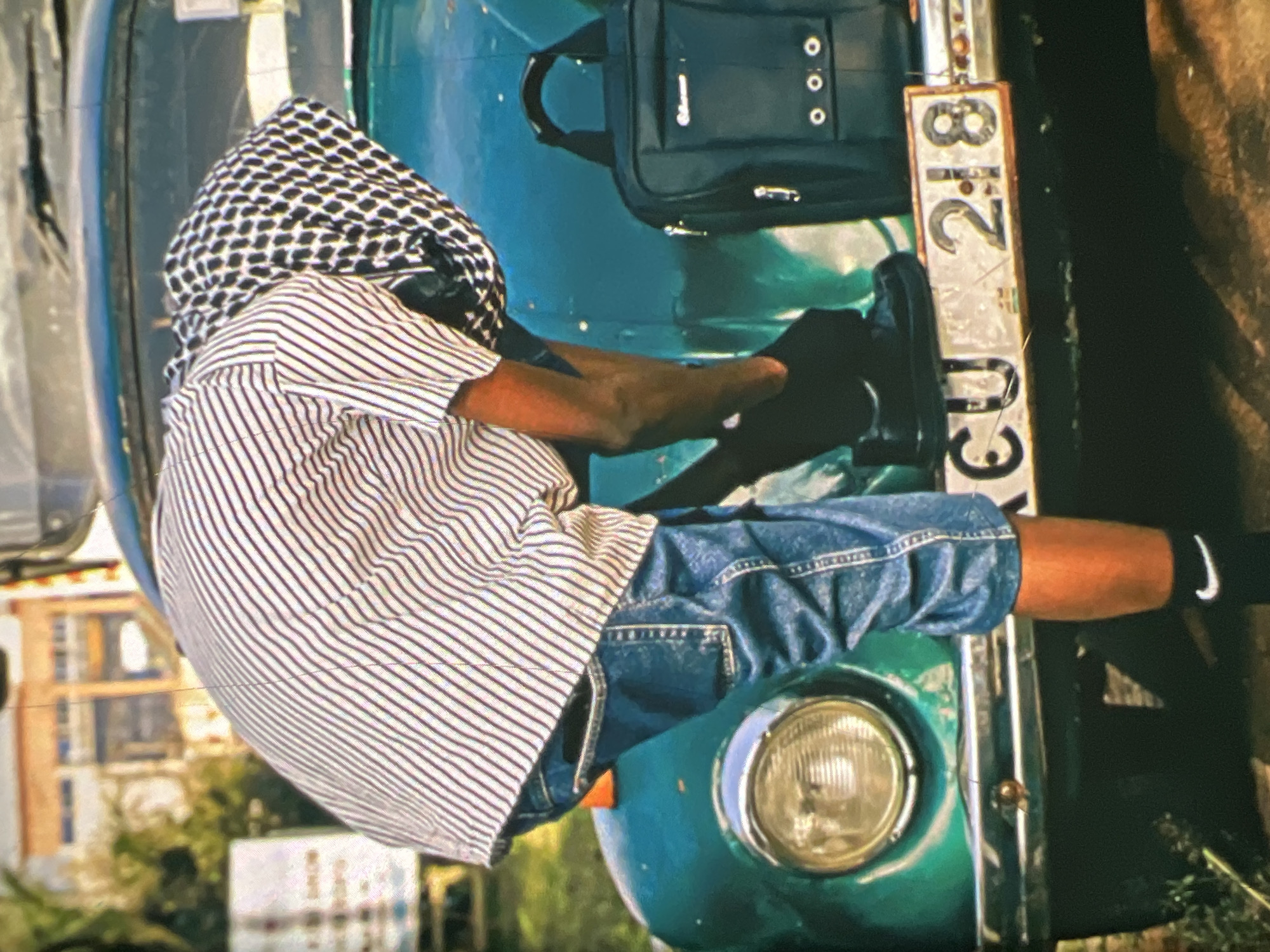 A person wearing a striped shirt, blue jeans, and black shoes is leaning over the front of a green vintage car in Lagos, Nigeria. The car has a visible license plate that reads "CU 218". A black bag is placed on the car's fender. The background shows a blurred building and some greenery. The person appears to be adjusting their shoe.