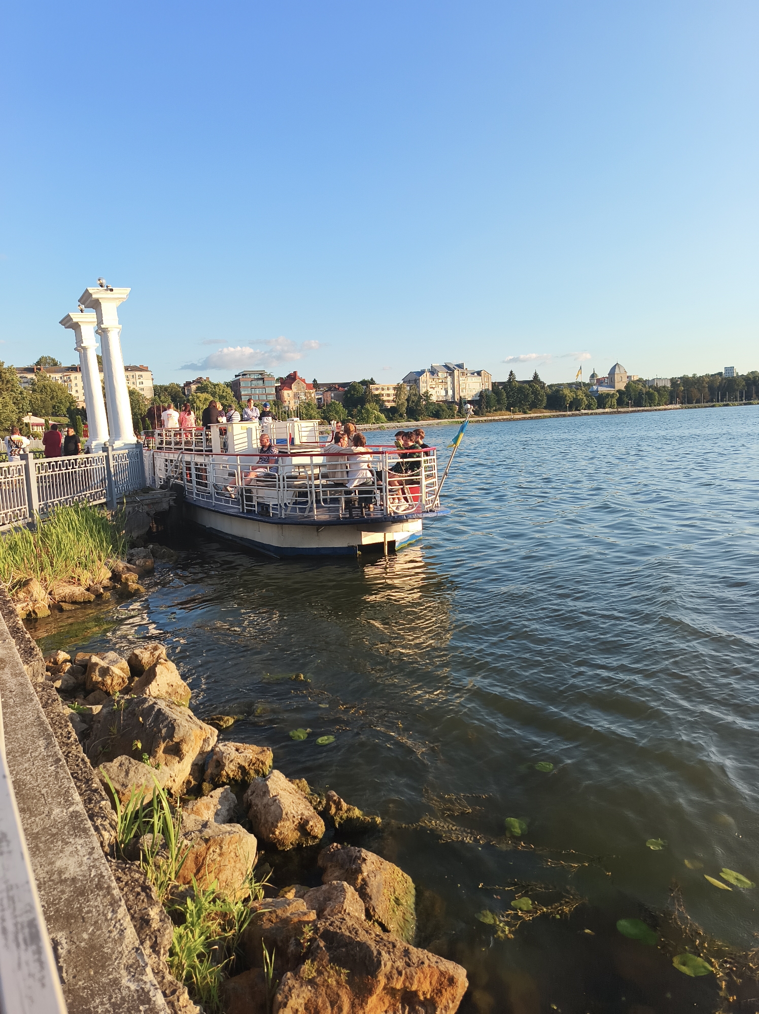 A view of Ternopil Lake in Ternopil, Ukraine, captured from the embankment on a clear day. The scene features a white passenger boat docked at the shore, with several people visible on its deck. The boat is white with red accents and a railing around the passenger area. In the background, a line of trees and buildings can be seen along the opposite shore, including what appears to be a domed structure. The water of the lake is calm with some ripples. The foreground includes a stone embankment with some vegetation and rocks near the water's edge. The sky is a clear blue, indicating pleasant weather.