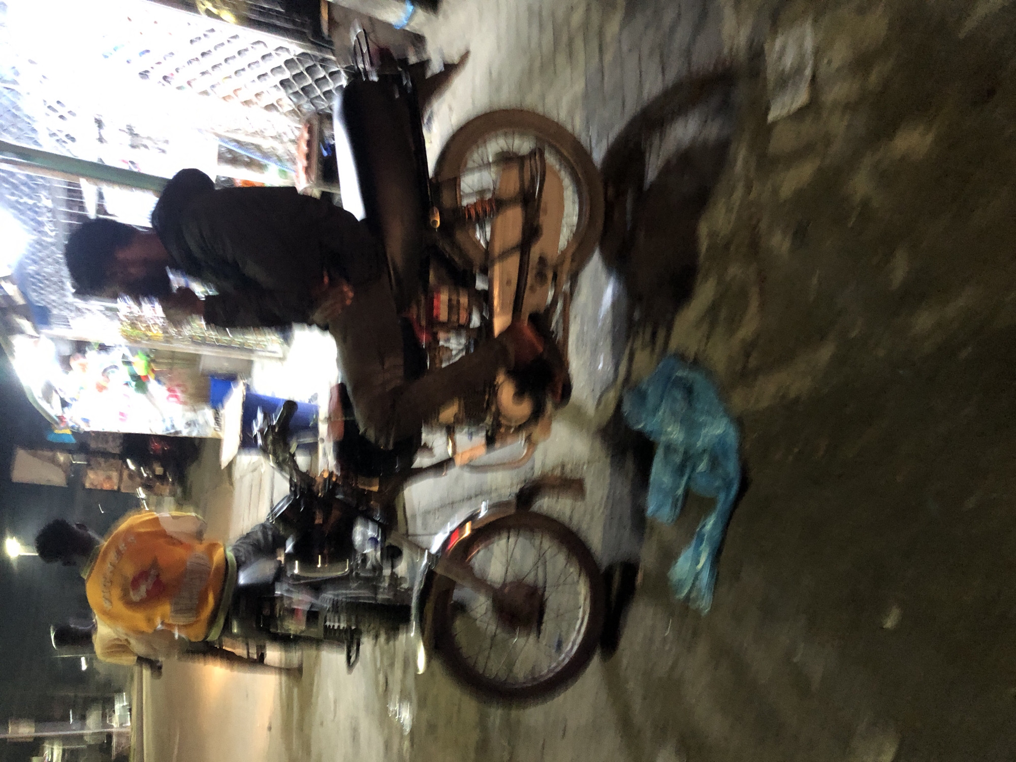 A blurry, low-light image shows two men with motorcycles on a street in Ado, Nigeria. One man in a dark jacket sits on a motorcycle near a shop with a brightly lit display. Another man wearing a yellow jacket sits on a second motorcycle further down the street. A blue plastic bag is on the ground. The street is dark and wet.