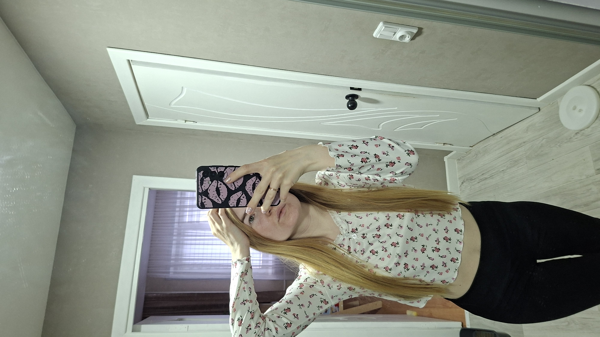 A fair-skinned, long-haired woman with blond hair takes a selfie in a mirror inside what appears to be a bathroom or hallway. She's wearing a floral-patterned long-sleeved crop top and black leggings. Her phone has a black and pink case with an abstract pattern. The reflection shows a white door, a light switch, and a window with blinds. Based on the image analysis, I can infer the city is in Chaikovsky, Russia. There's no specific geographical or landmark information directly visible to confirm the location inside the city.