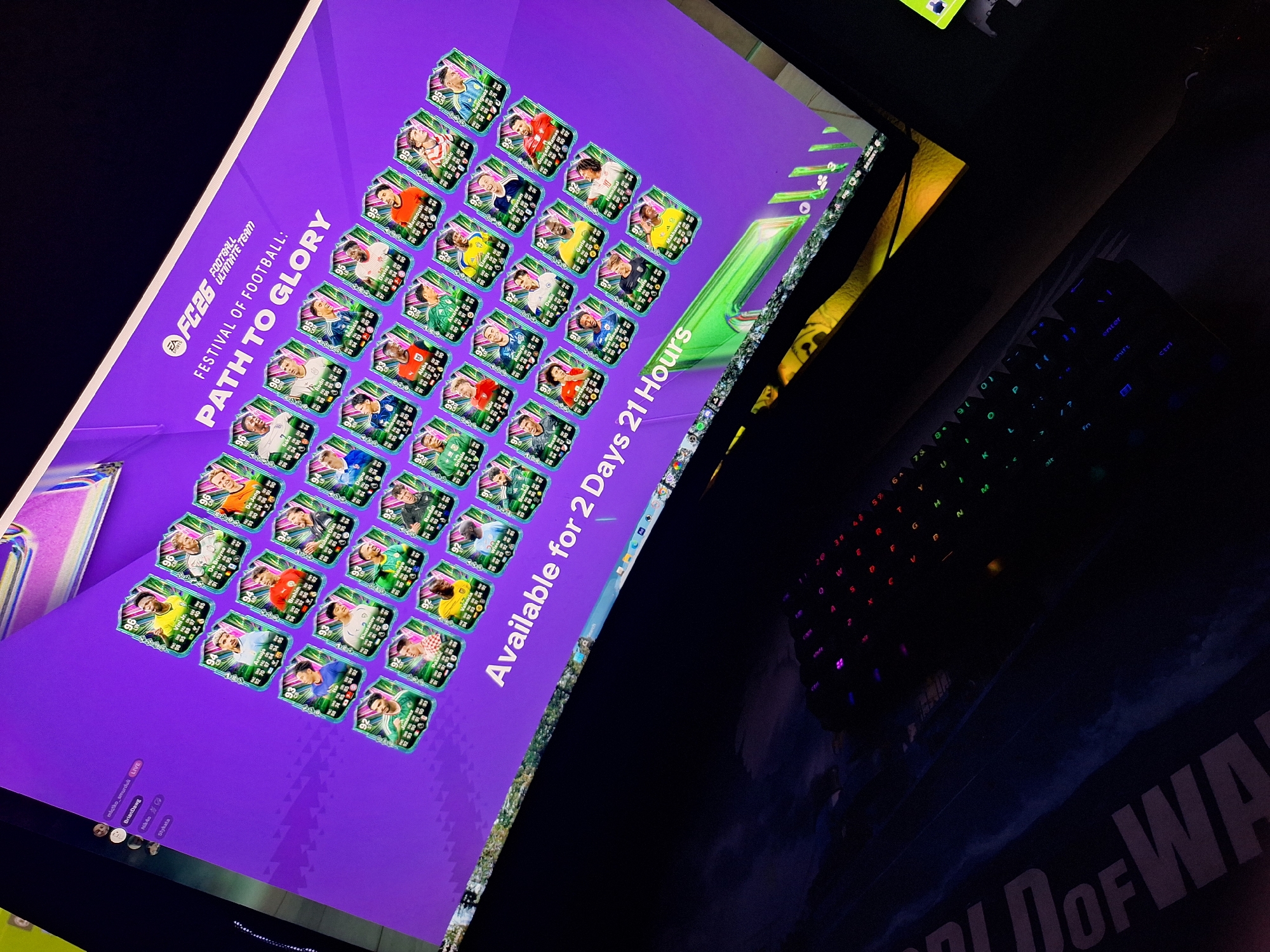 This is a dimly lit, high-angle photograph of a computer monitor displaying the "FC 24 Football Ultimate Team: Festival of Football: Path to Glory" interface. The screen shows numerous player cards, each with a player's image, rating, and stats. The background of the interface is purple. Below the monitor, a black mechanical keyboard with illuminated keys is visible. The lower portion of the image shows a dark surface with a partially visible graphic that appears to be from the "Call of Duty: Modern Warfare" franchise. There is no discernible information about the specific location within the city of Varna, Bulgaria. No people are visible in the frame.