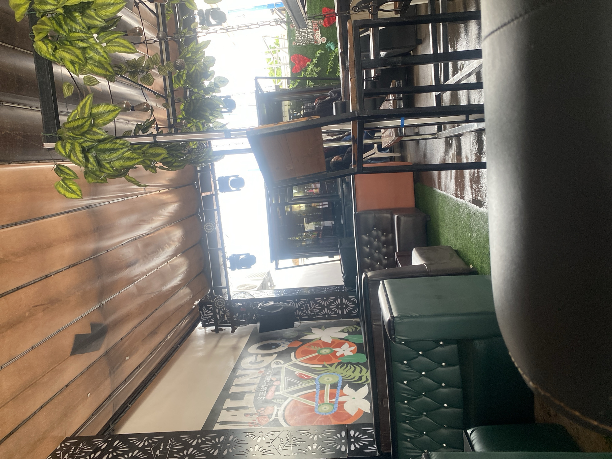 This image depicts a semi-outdoor establishment, likely a restaurant or bar, located in Howayi, Nigeria. The scene is illuminated by ambient light, suggesting daytime or a brightly lit evening.
The ceiling consists of a light brown, possibly fabric or wooden, retractable awning or canopy structure supported by metal tracks. Several dark marks and reflections are visible on its surface. Artificial green leafy vines hang from black metal support beams throughout the overhead area, interspersed with hanging light fixtures resembling Edison bulbs. Additional stage-style spotlights are mounted on metal trusses.
The left wall features a large mural. The mural prominently displays the word "ALINGOO" above "STEADY CHILLING," accompanied by a stylized bicycle with oversized orange wheels, a blue chain, white flowers, and green leaves. Adjacent to this mural, and recurring as a decorative element, are black metal screens with intricate cut-out patterns.
The seating arrangements include deep green, tufted booth seating with diamond patterns and silver stud accents, as well as darker brown or black upholstered armchairs. The floor alternates between artificial green turf and a darker surface, possibly concrete or tile, which appears wet or reflective in several areas.
In the middle-ground, a tall, dark metal-framed chair with a wooden back partially obstructs the view. The foreground features an out-of-focus, dark-colored upholstered armrest.
Towards the right side of the image, a dark bar or counter area is visible. Behind it, a green wall decor features two large red heart shapes made of artificial foliage and a white sign that reads "OUR BE IS COLD THAN YOUR EX-HURT," likely intending "OUR BEER IS COLDER THAN YOUR EX-HEART." White air conditioning units are mounted on a wall in the far background. Several indistinct individuals are visible in the background, seated or standing within the establishment.