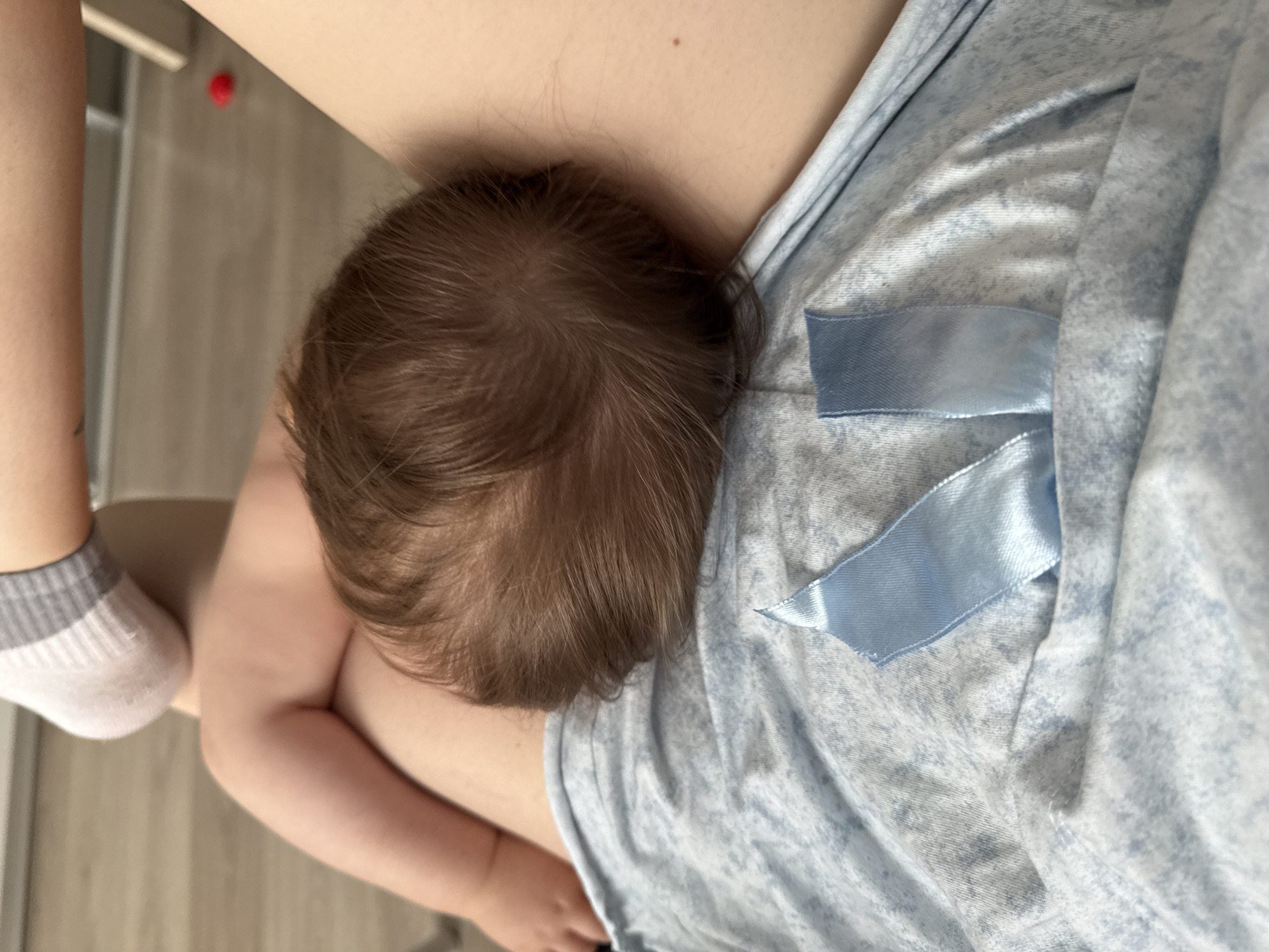 A baby with brown hair is resting its head against an adult's chest. The adult is wearing a light blue patterned garment with a blue ribbon. The adult has a white and gray sock on one foot. The background is a light-colored floor. The image does not appear to be in Moscow, Russia.